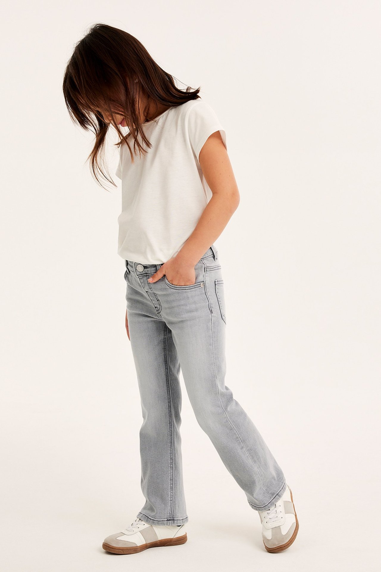 Sivukuva lapsesta harmaissa denim bootcut-farkuissa painonapilla, valkoisessa t-paidassa ja tennareissa.