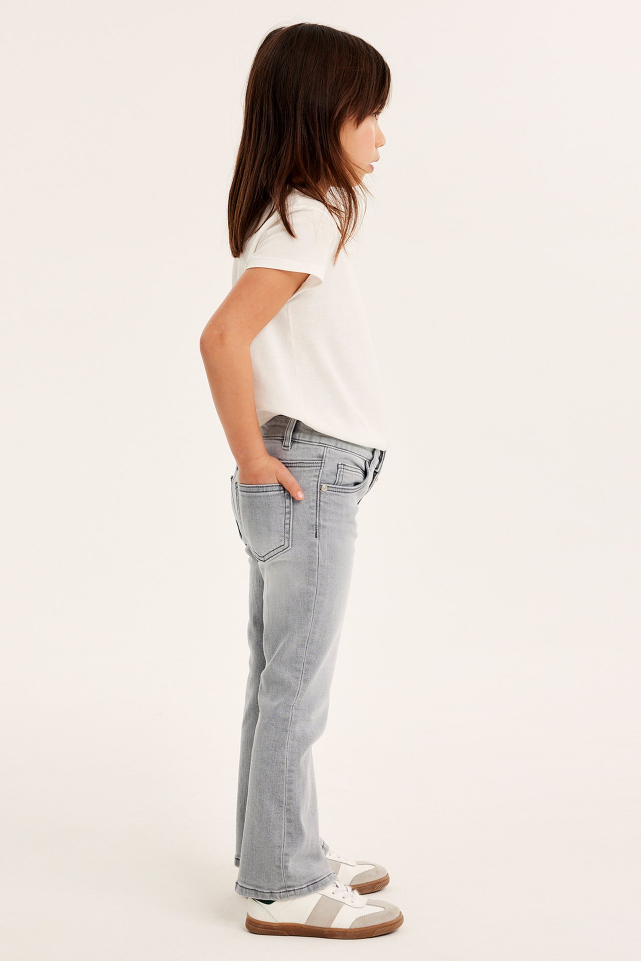 Sidovy av ett barn i grå bootcut-jeans med medelhög midja, en vit t-shirt och vita/beige sneakers.