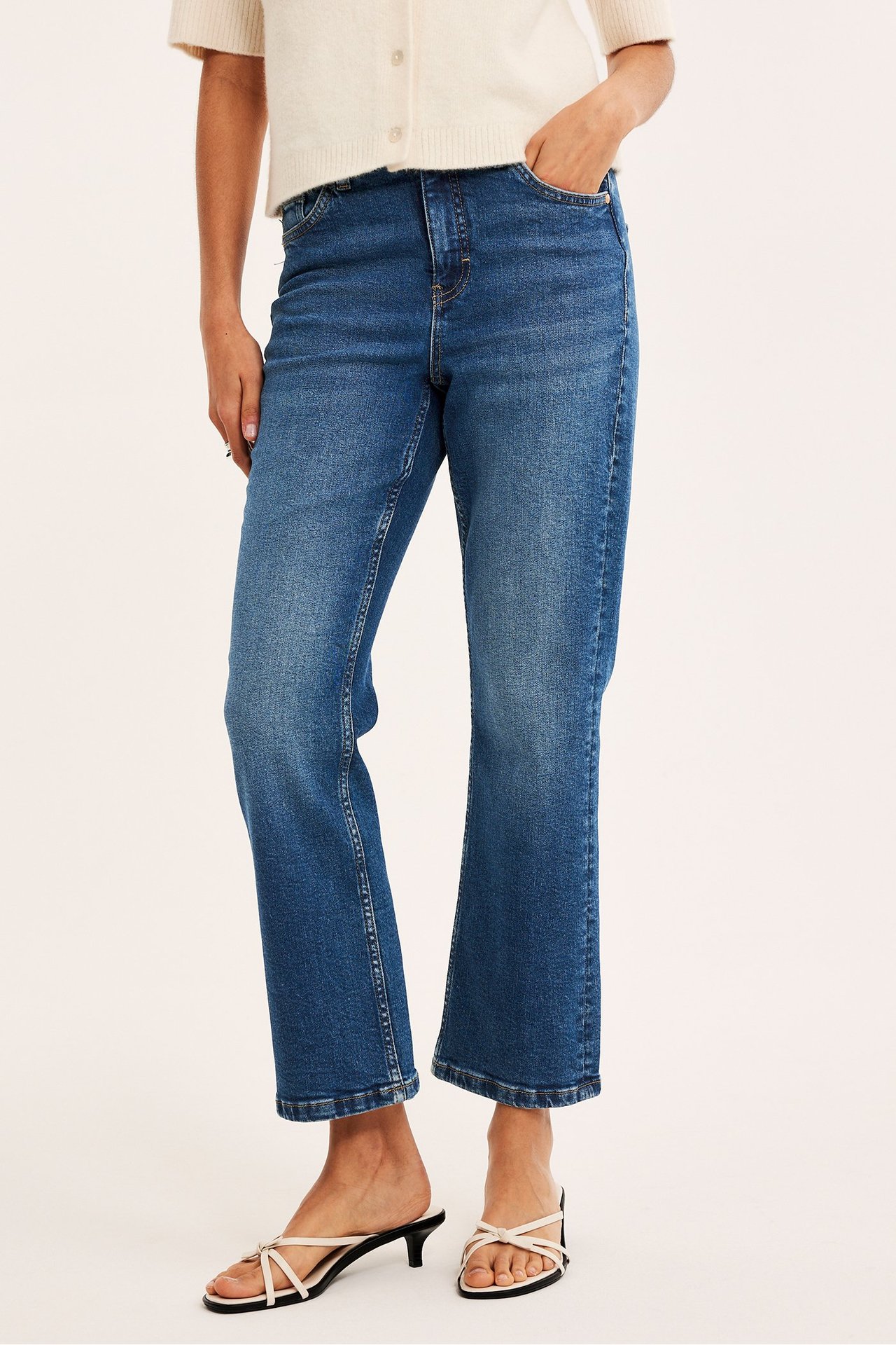Framifrån: kvinna i blå denim cropped flare jeans, ljusbeige kofta och vita sandaler.