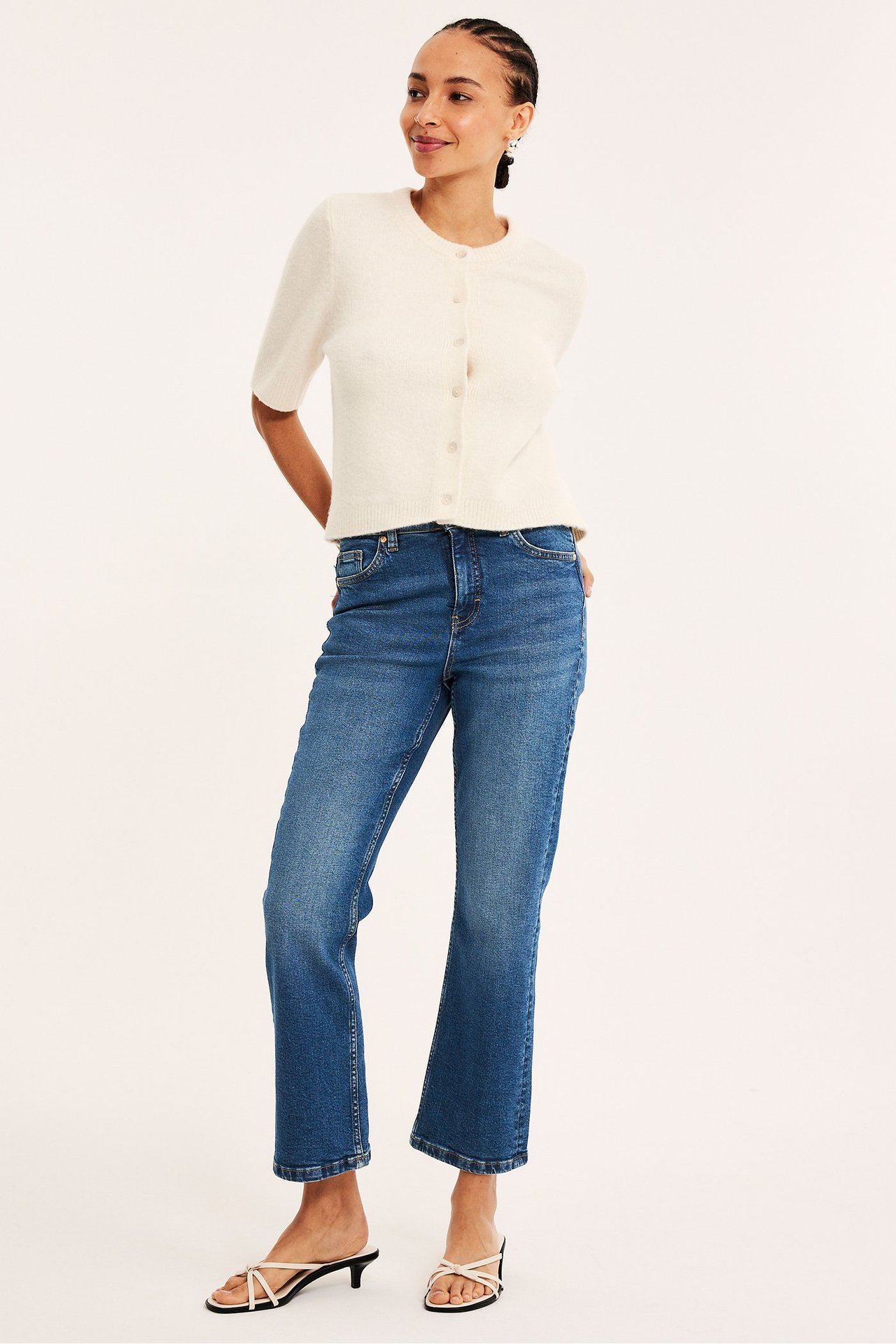 Framifrån: kvinna i blå cropped flare jeans, ljusbeige kortärmad kofta och vita remsandaler.