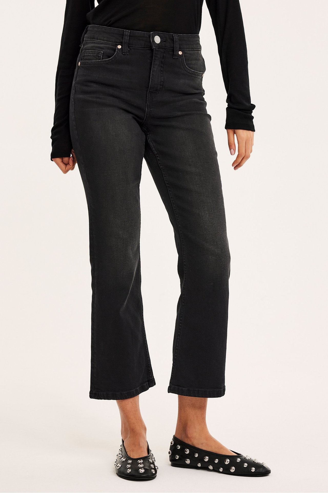Framifrån: kvinna i ljussvarta cropped flare jeans, svart topp och svarta nitprydda ballerinaskor.