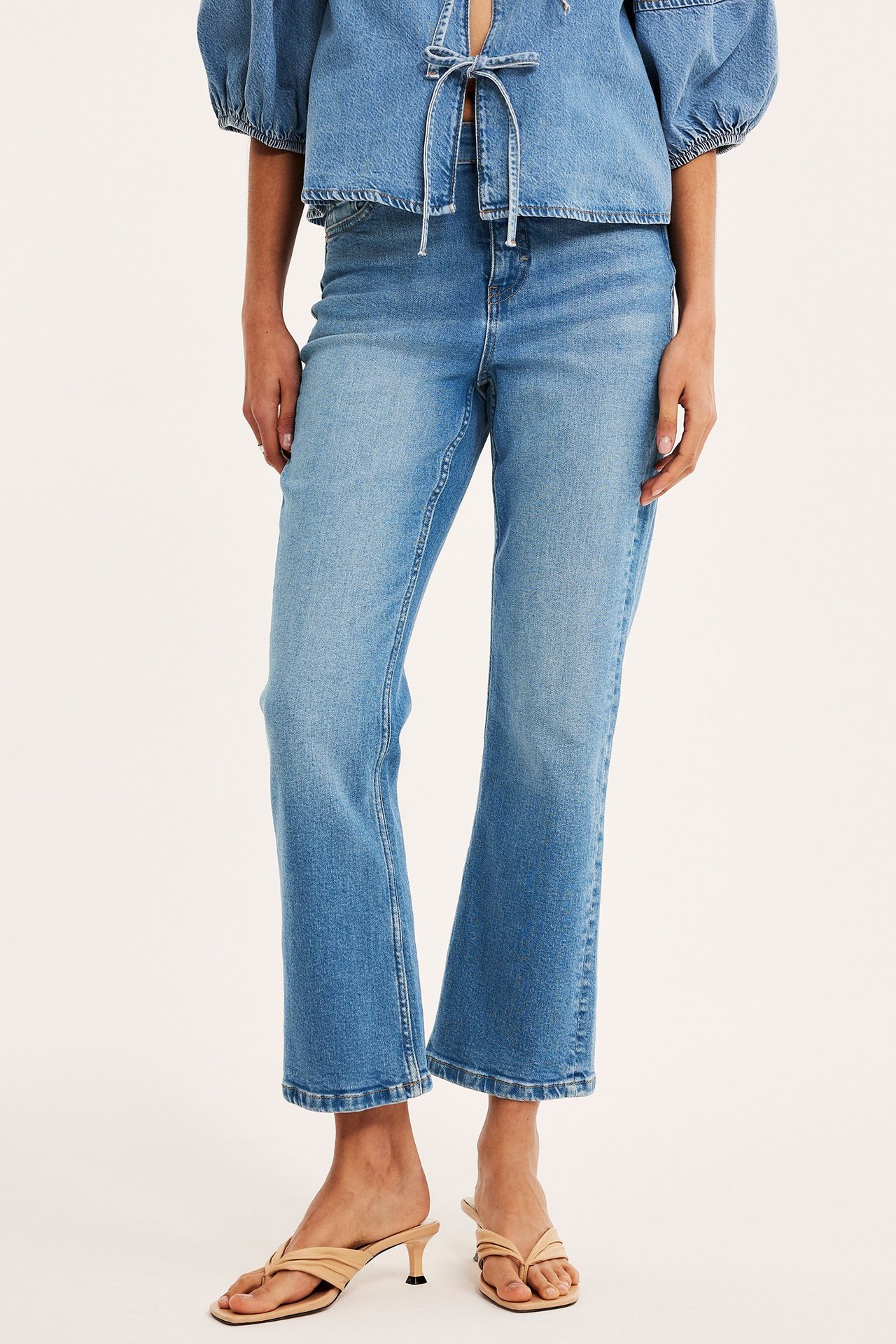 Framifrån: Kvinna i ljusblå cropped flare jeans, en denimtopp med knytning och beiga klacksandaler.