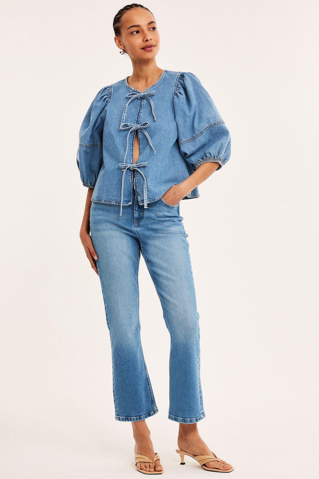 Framifrån: Kvinna i ljusblå cropped flare jeans, en denimtopp med rosetter och beiga klacksandaler.