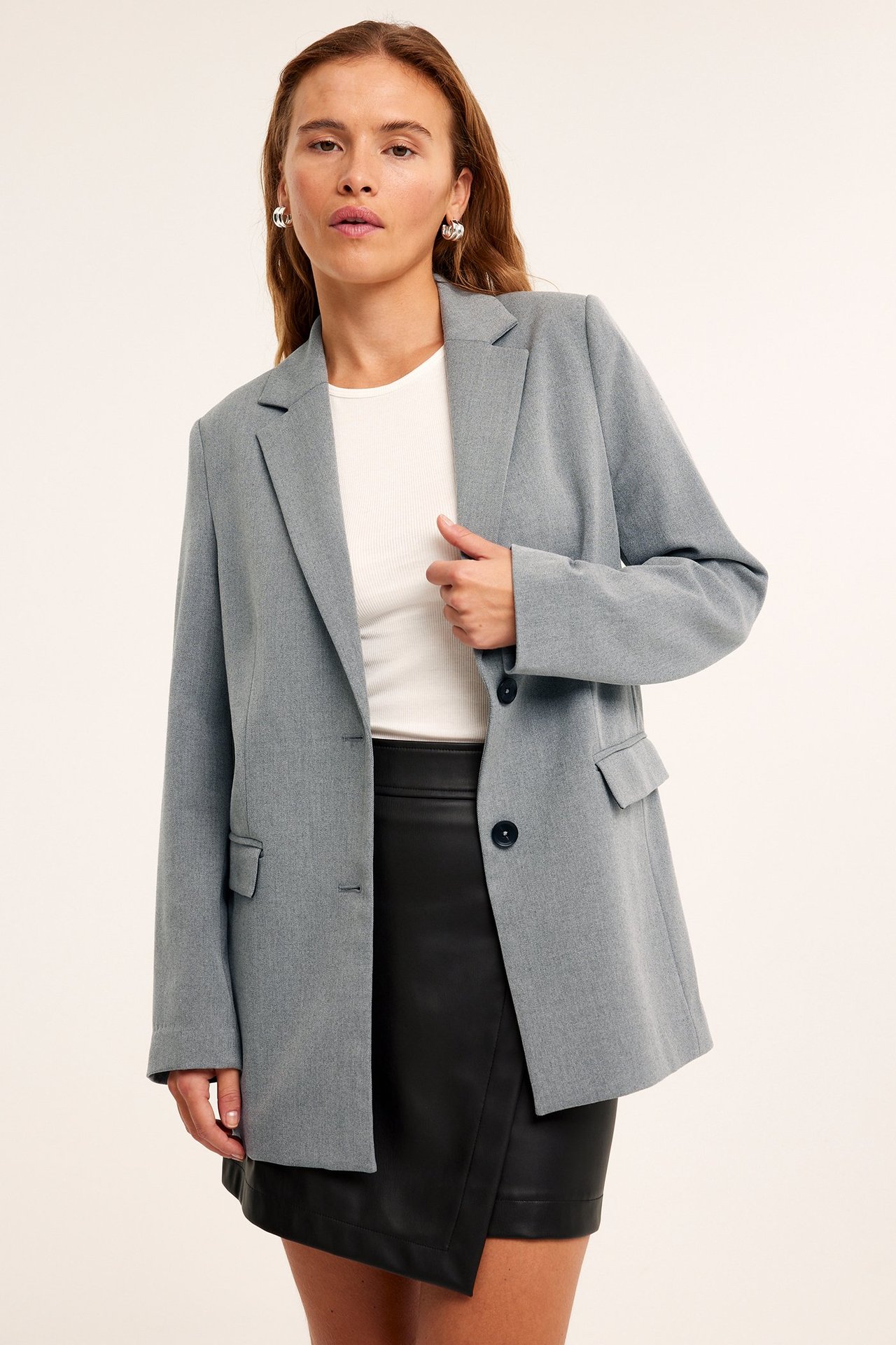 Forfra, kvinne i lys grå melert oversized blazer, hvit ribbet topp og svart skjørt i imitert skinn.
