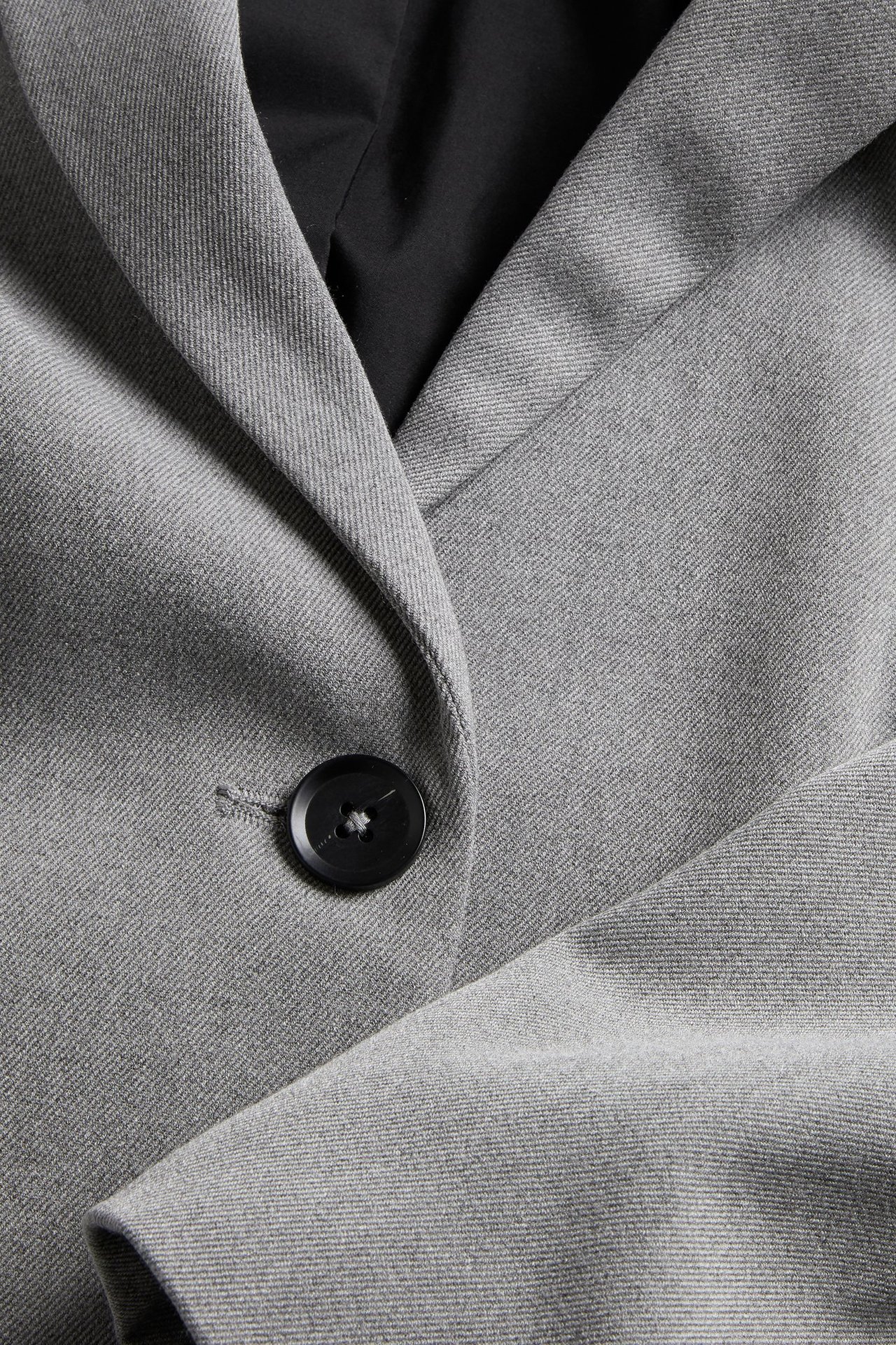 Zbliżenie jasnoszarej melanżowej marynarki oversize z czarnym guzikiem i czarną podszewką.