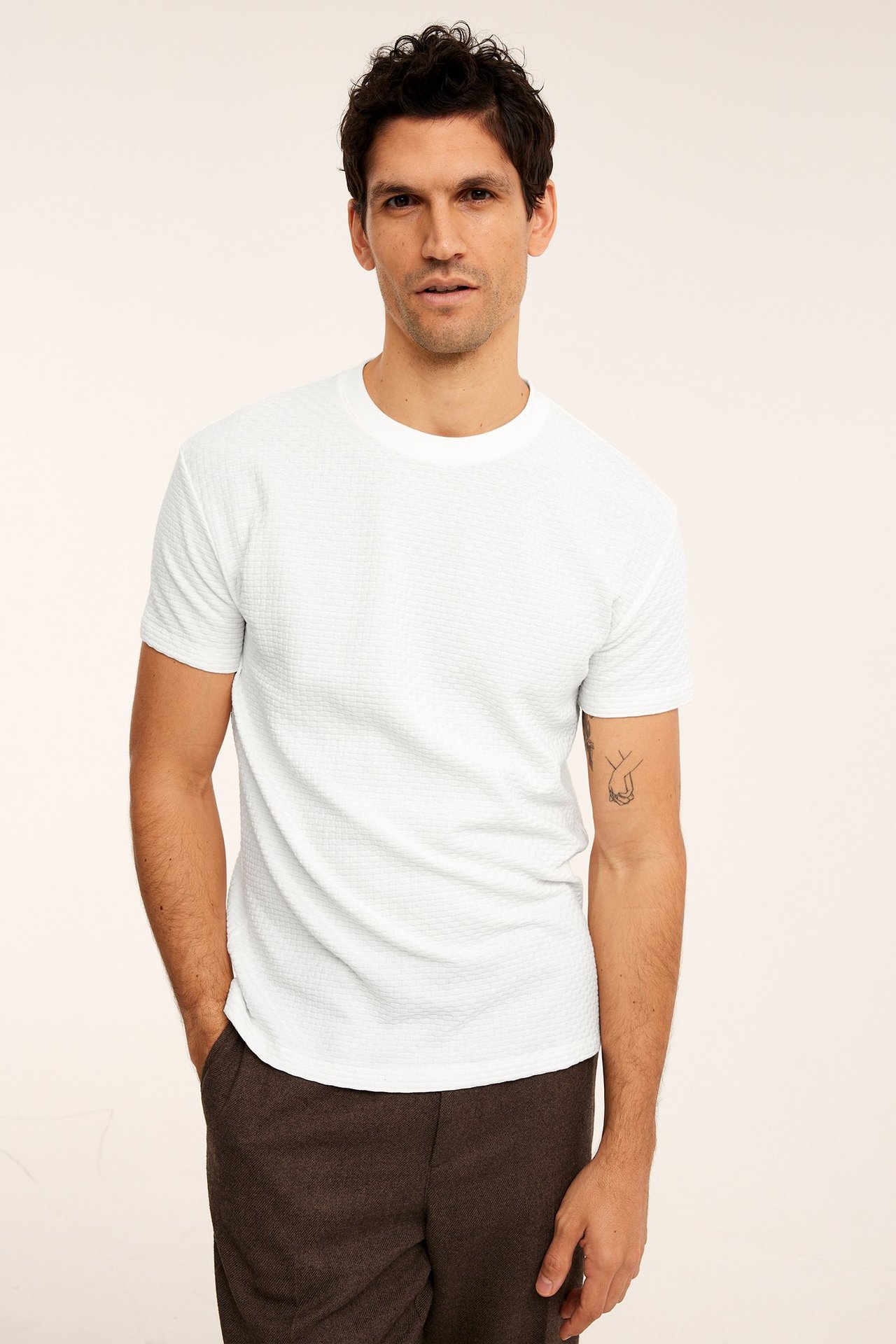 Framifrån, man i offwhite strukturerad bomulls-T-shirt och mörkbruna byxor, hand i fickan.