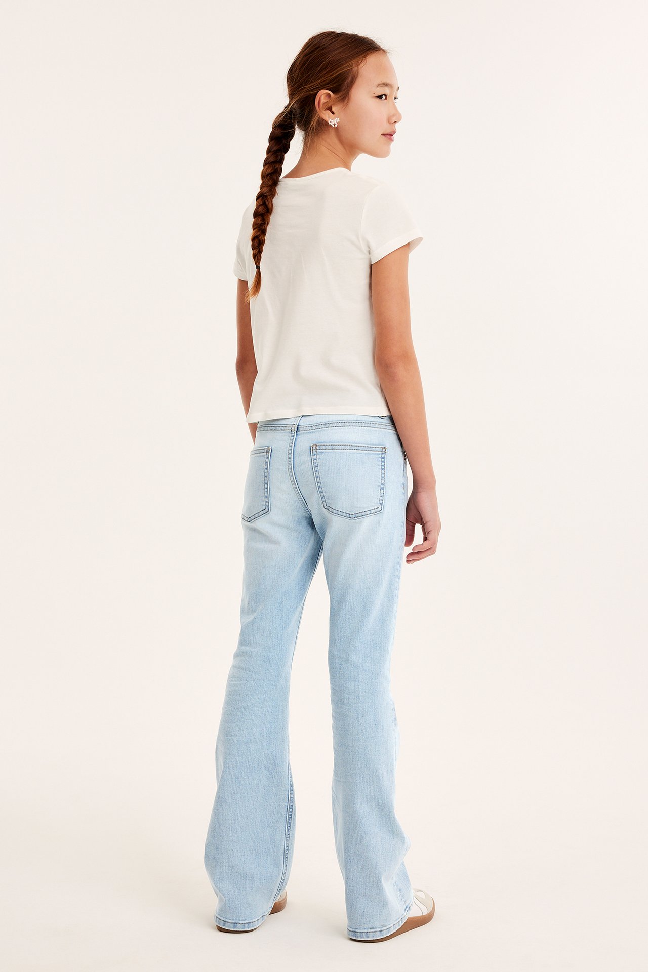 Widok z tyłu dziecka w jasnych dżinsach bootcut, białym t-shircie i jasnych butach.