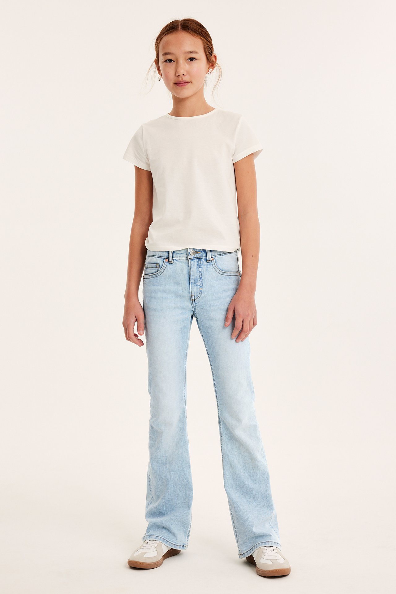 Widok z przodu: dziecko w jasnoniebieskich jeansach bootcut z wysokim stanem, białym T-shircie i jasnych sneakersach.