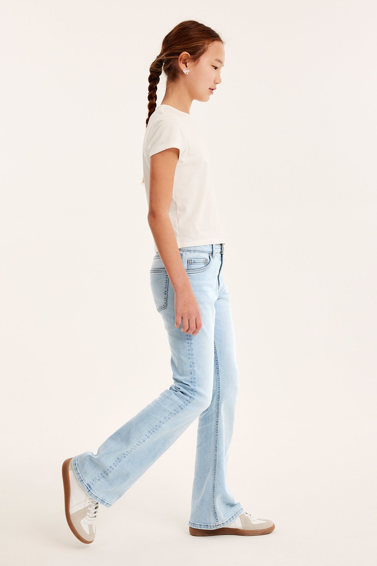 Widok z boku: dziecko w jasnoniebieskich jeansach bootcut, białym t-shircie, białych-beżowych sneakersach i kolczykach.