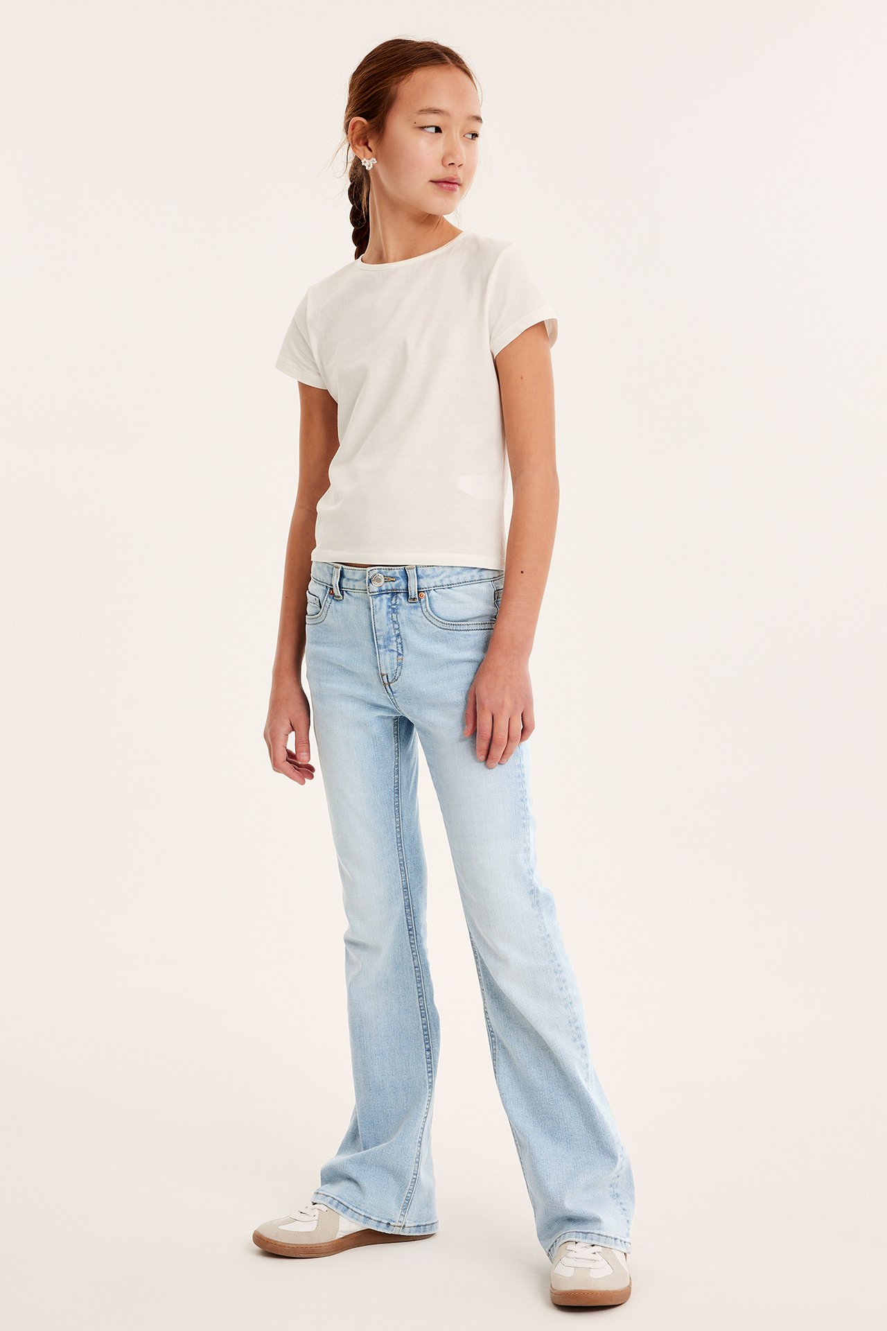 Widok z przodu dziecka w jasnoniebieskich jeansach bootcut, białym t-shircie i jasnych sneakersach.