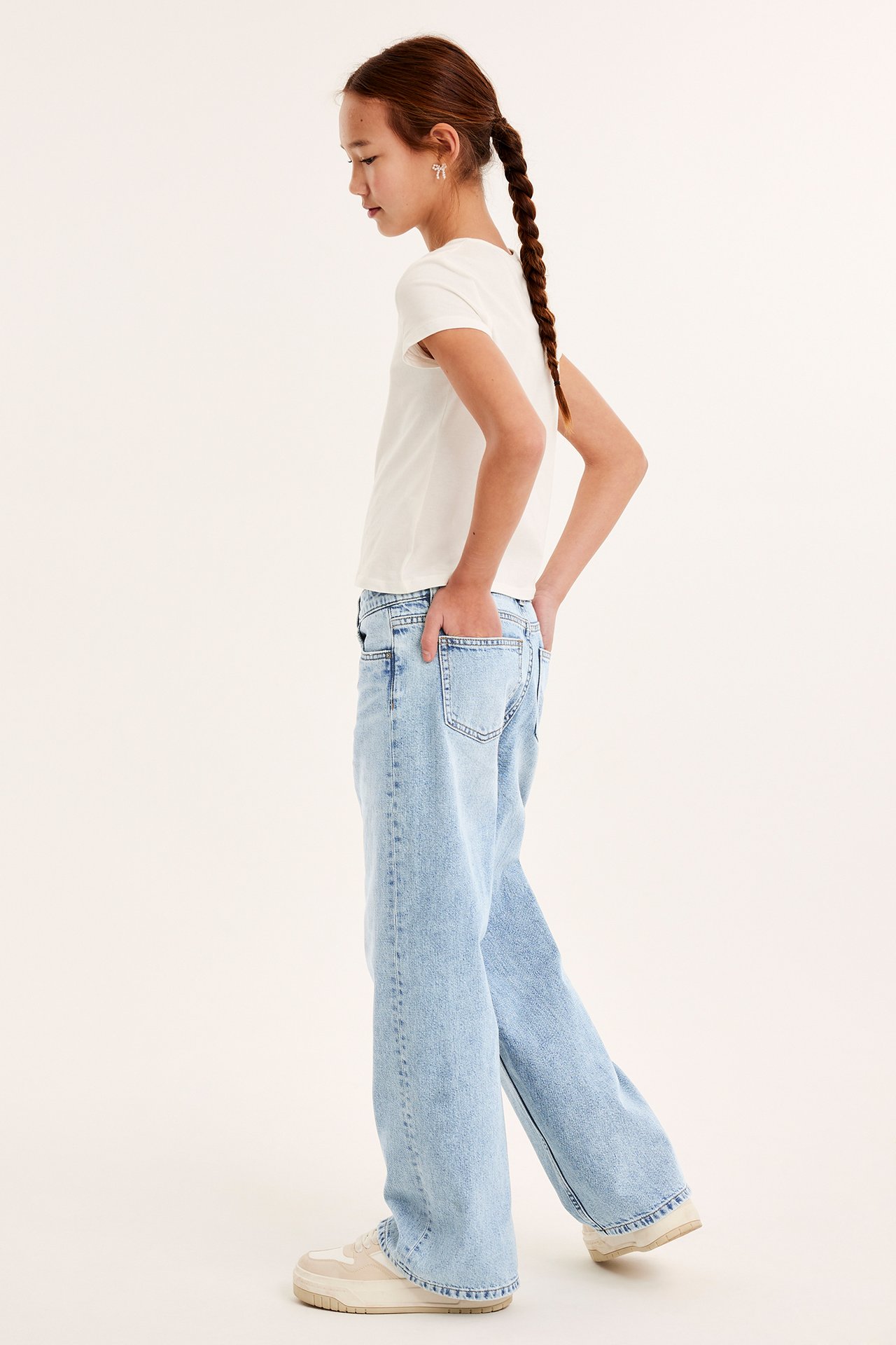 Sidebilde av et barn i lyse løse lavtaljede jeans, en hvit t-skjorte og beige joggesko.