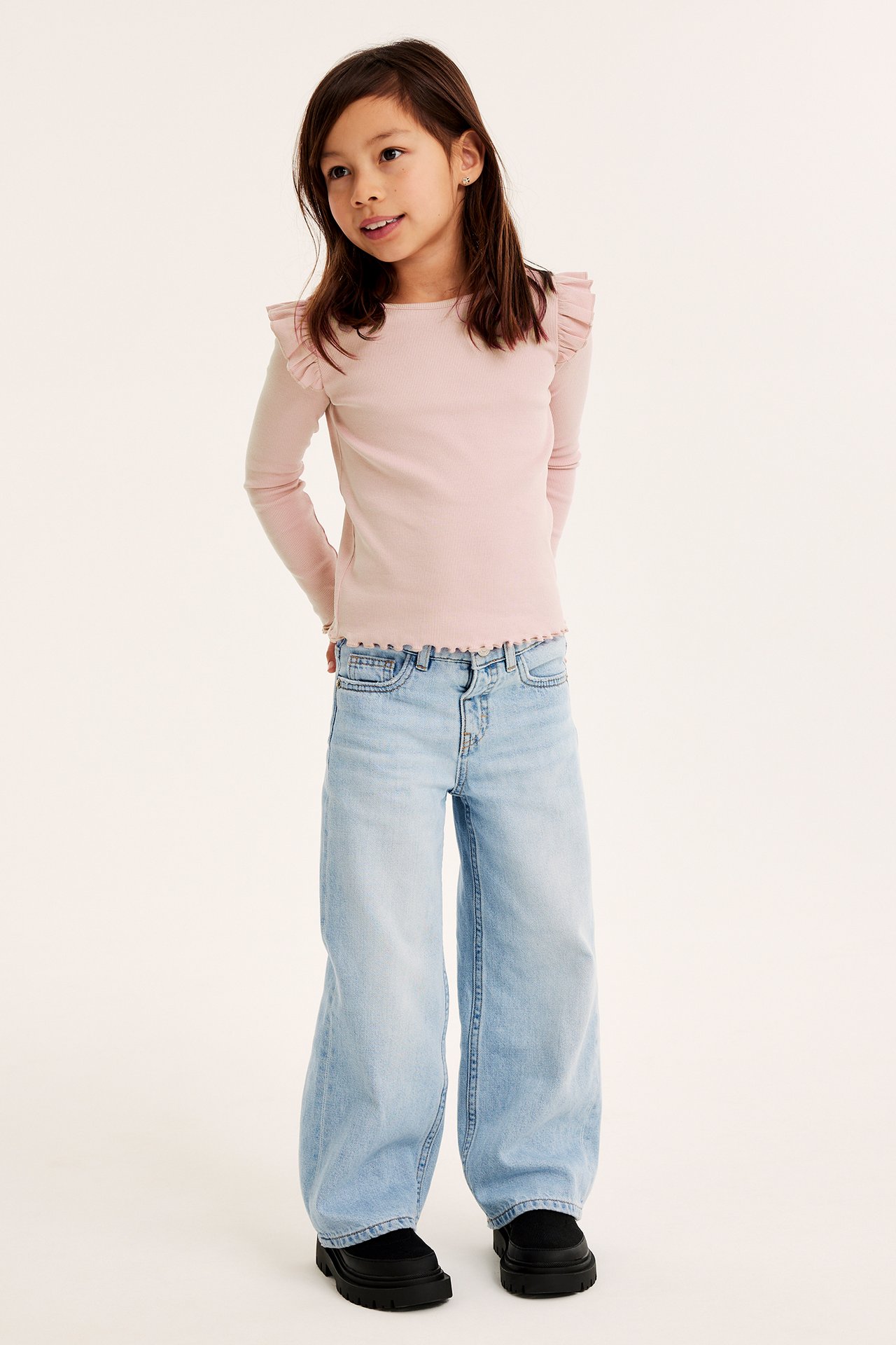 Forfra: Barn i lys rosa ribbet volangtopp, lyseblå vide jeans og svarte chunky sko.