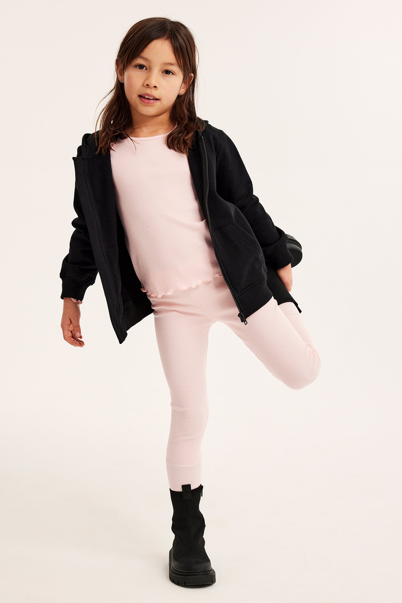 Framifrån: barn i ljusrosa ribbade leggings, matchande topp, svart huvtröja med dragkedja och svarta kängor.
