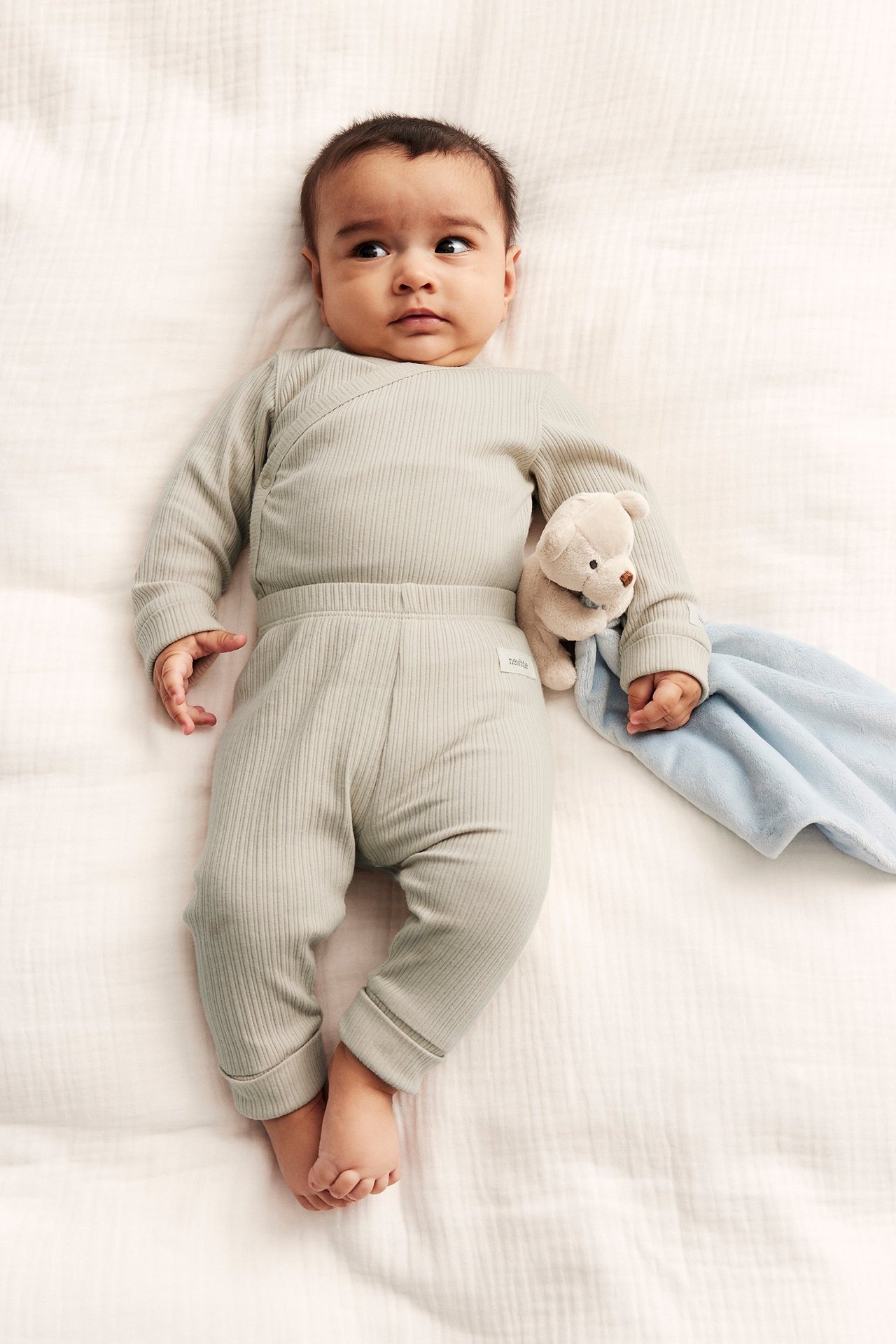 Ovanifrån: baby i beige ribbad omlottbody, matchande byxor, håller nallebjörn och blå filt.