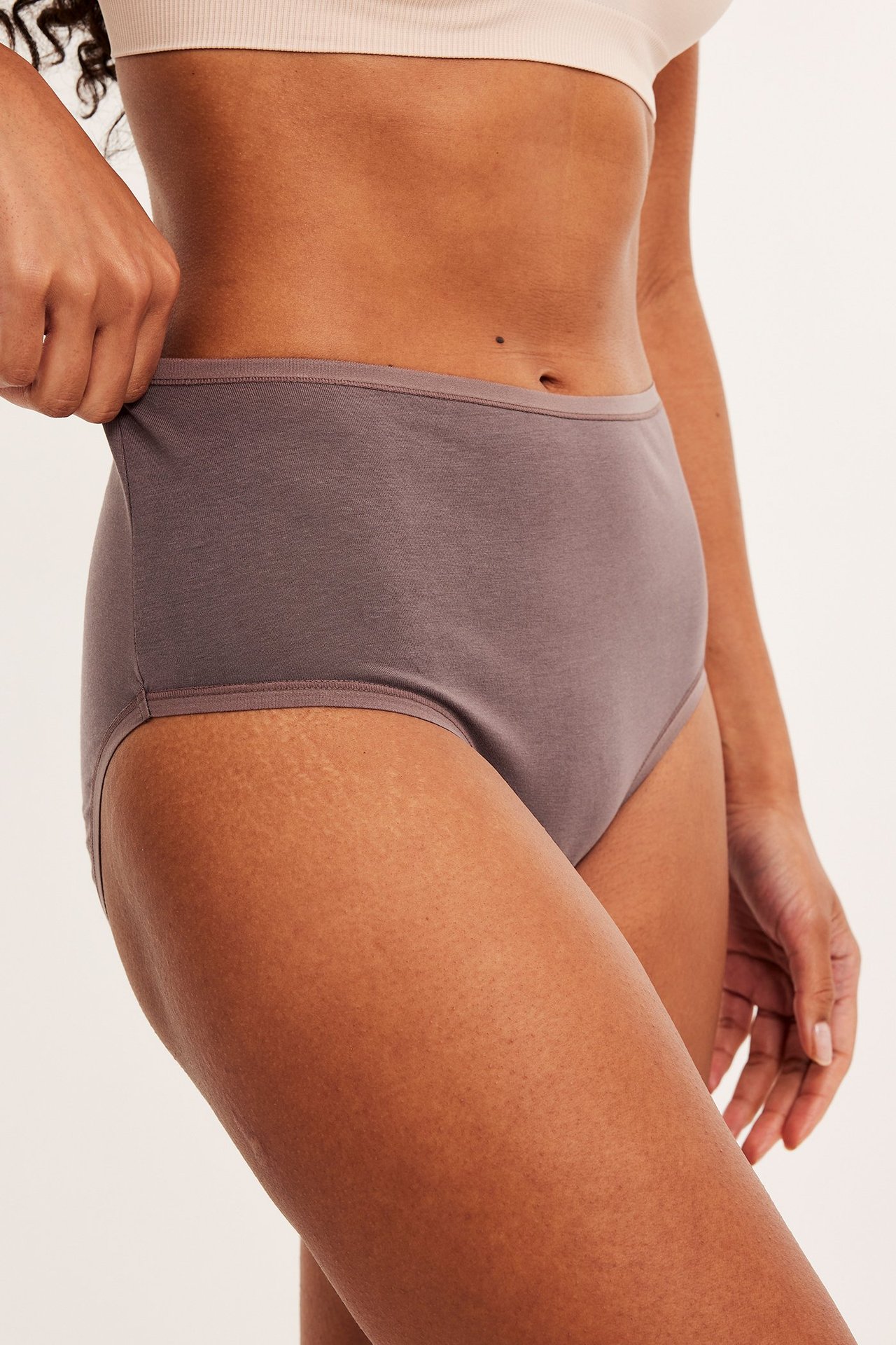 Sidovy av kvinna i plommonfärgade högmidjade bomullstrosor och en ljusbeige bralette.