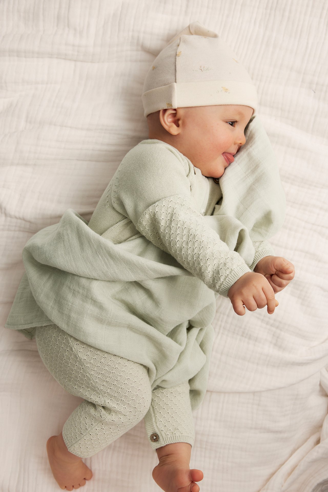 Ovenfra: baby i lys grønn strikket jumpsuit med tekstur, beige lue og grønt teppe.