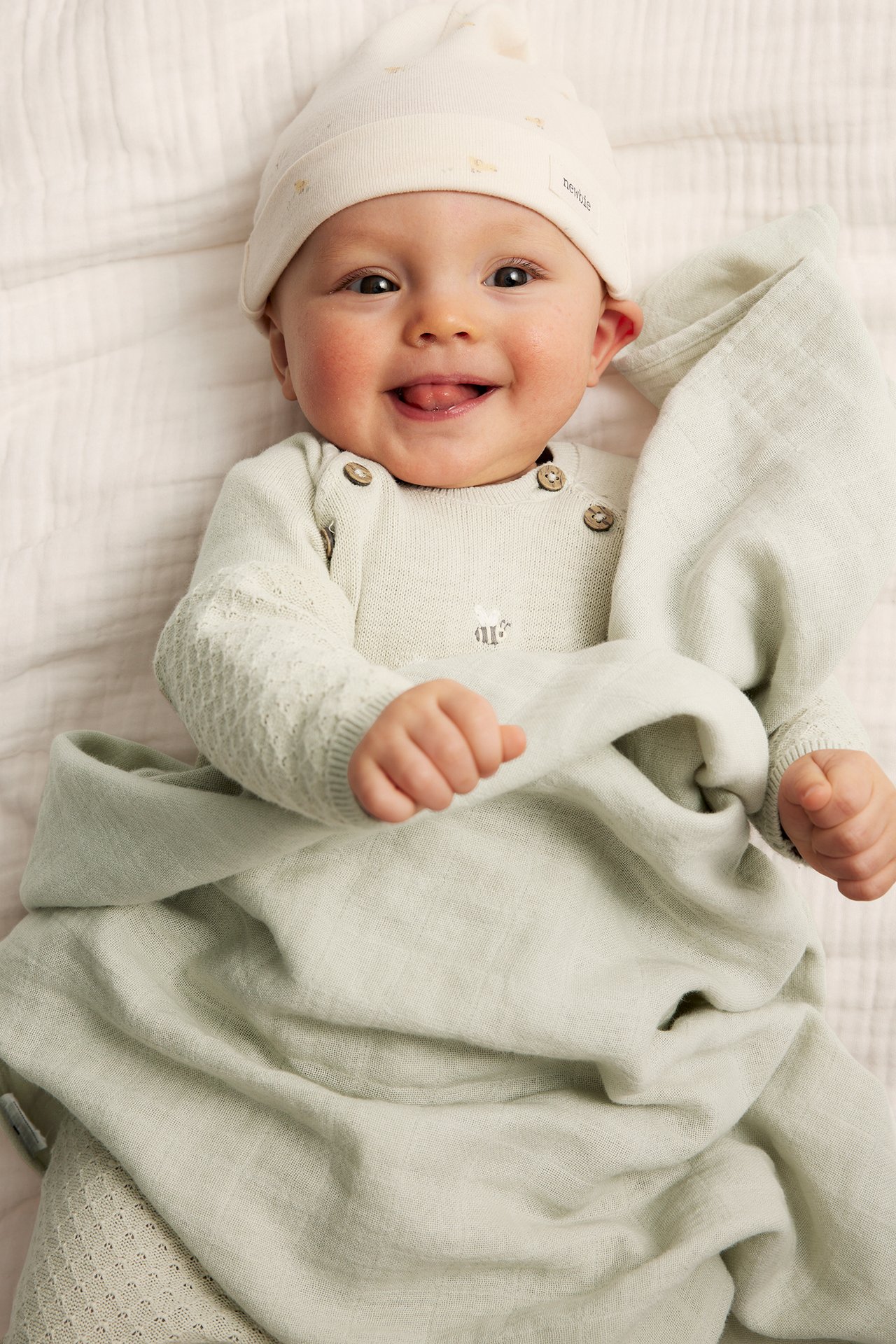 Smilende baby i lysegrønn strikket bomullsjumpsuit med knapper, broderi og en kremfarget lue.