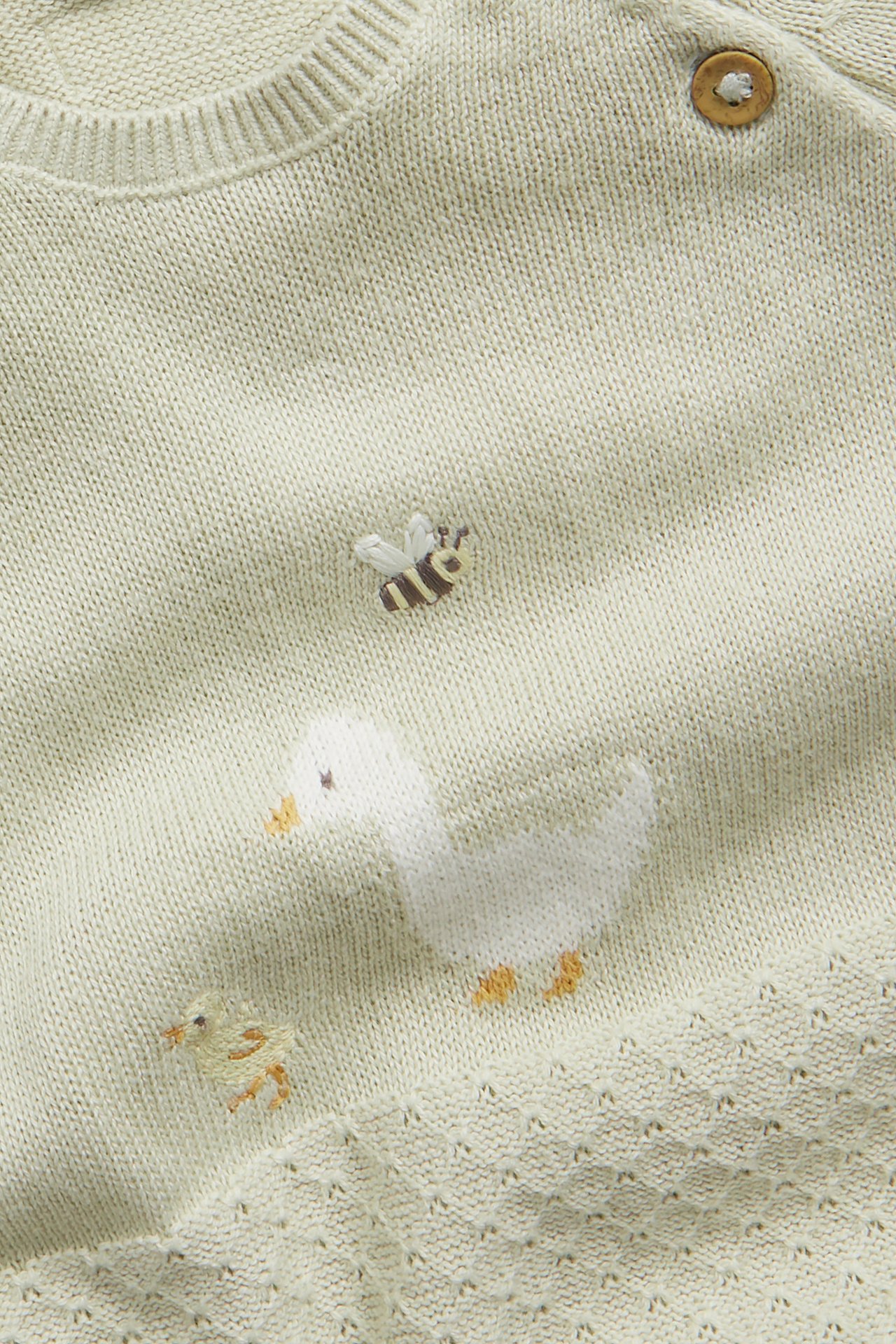 Nærbilde av lys grønn strikket babyjumpsuit med broderte and, kylling og bie, og en treknapp.