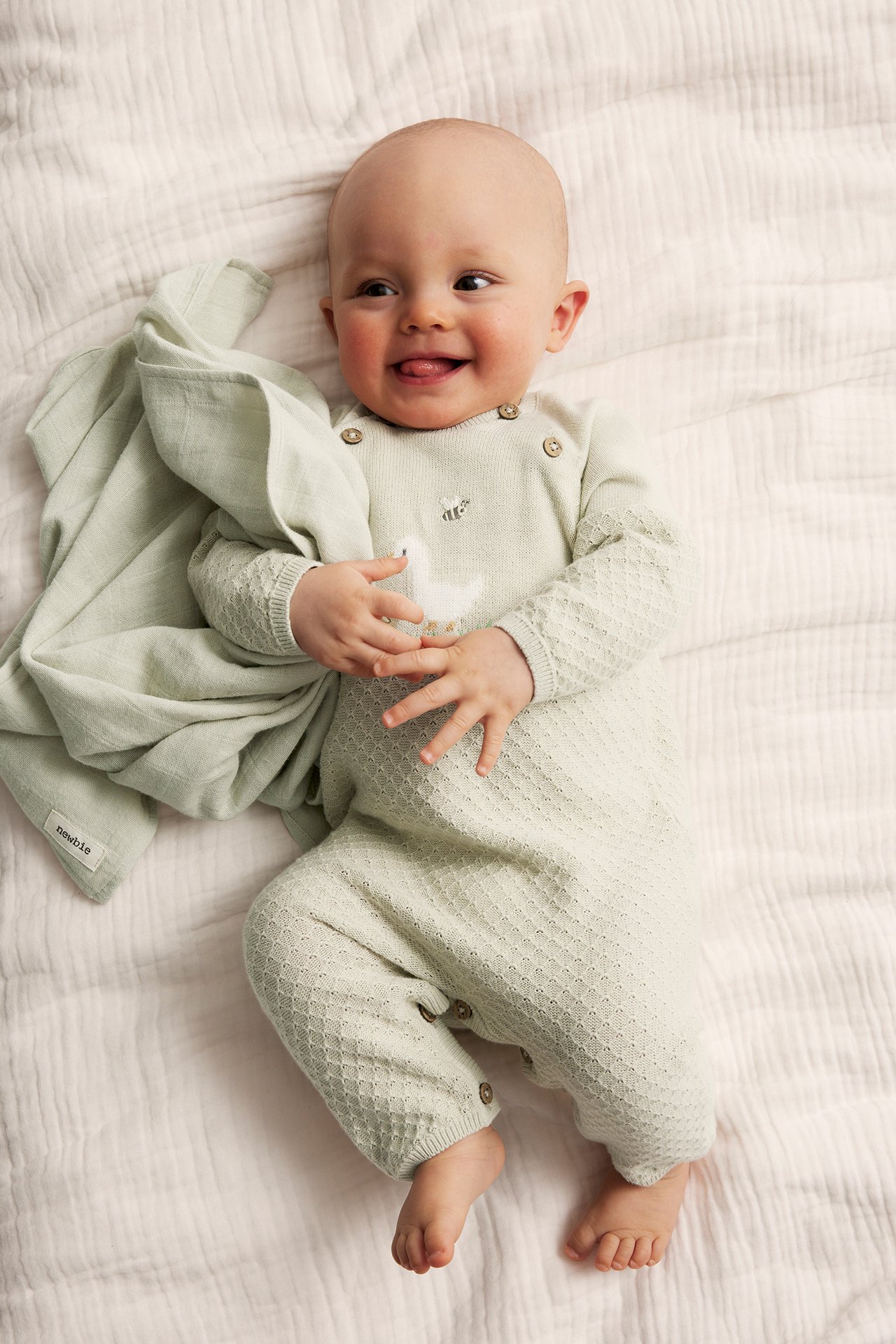 Ovenfra-visning av en smilende baby i lysegrønn strikket jumpsuit med andebroderi, holder et matchende teppe.