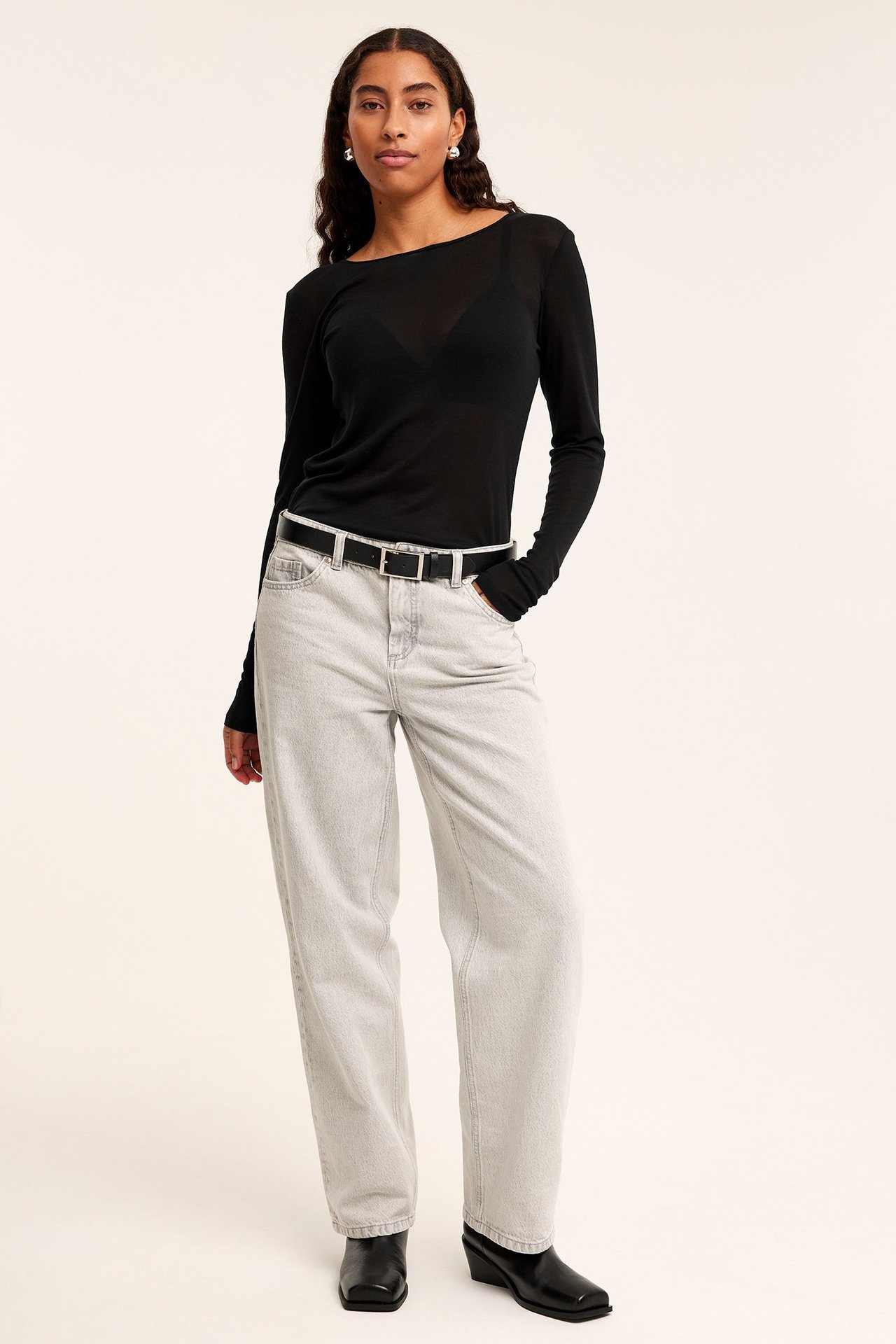 Frontbilde: Kvinne i gjennomsiktig svart langermet tenceltopp, lysegrå jeans, svart belte og svarte støvler.