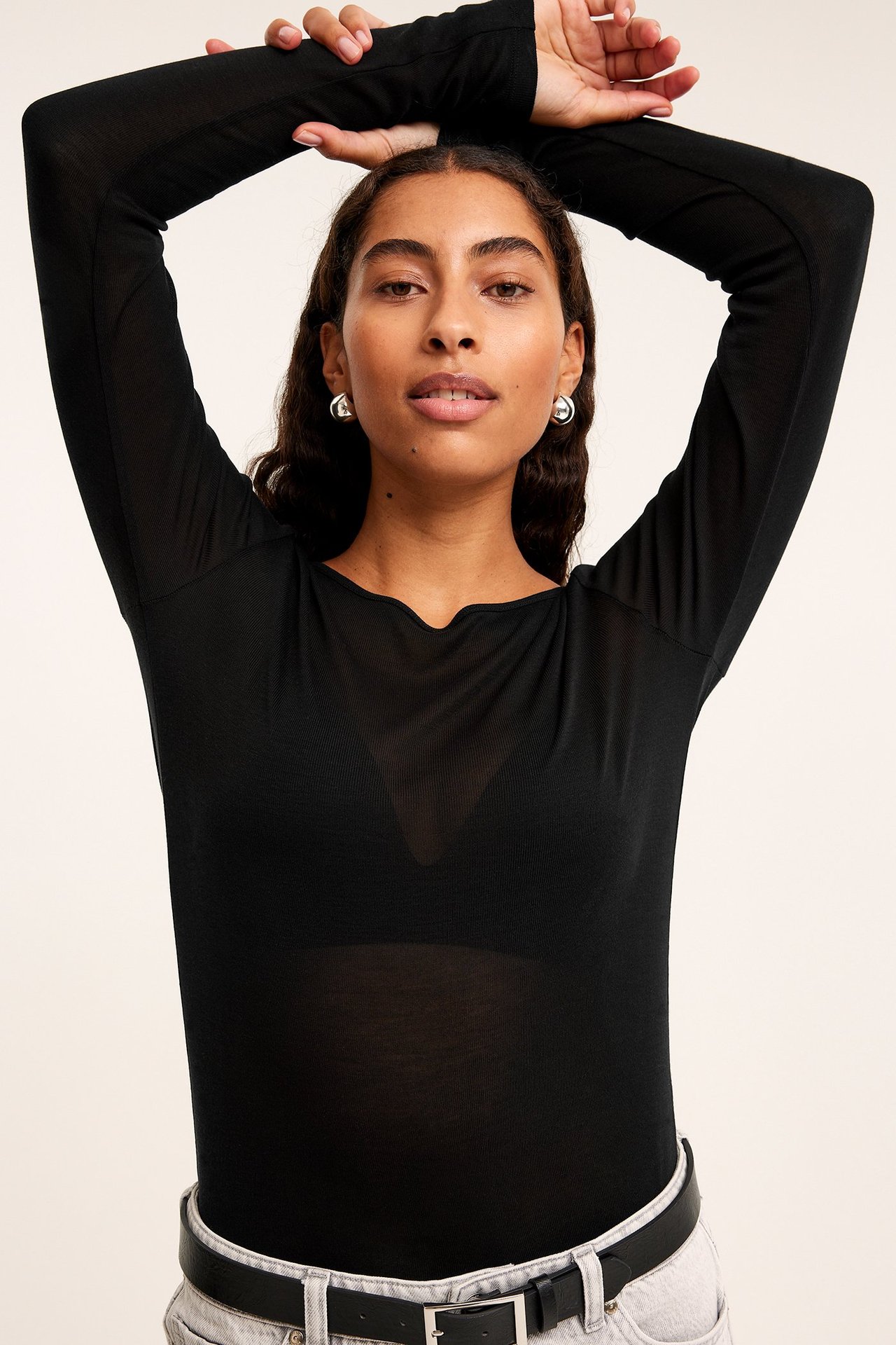 Frontvisning: kvinne i en tynn svart langermet lyocell-topp, svart BH, lyse jeans og belte.