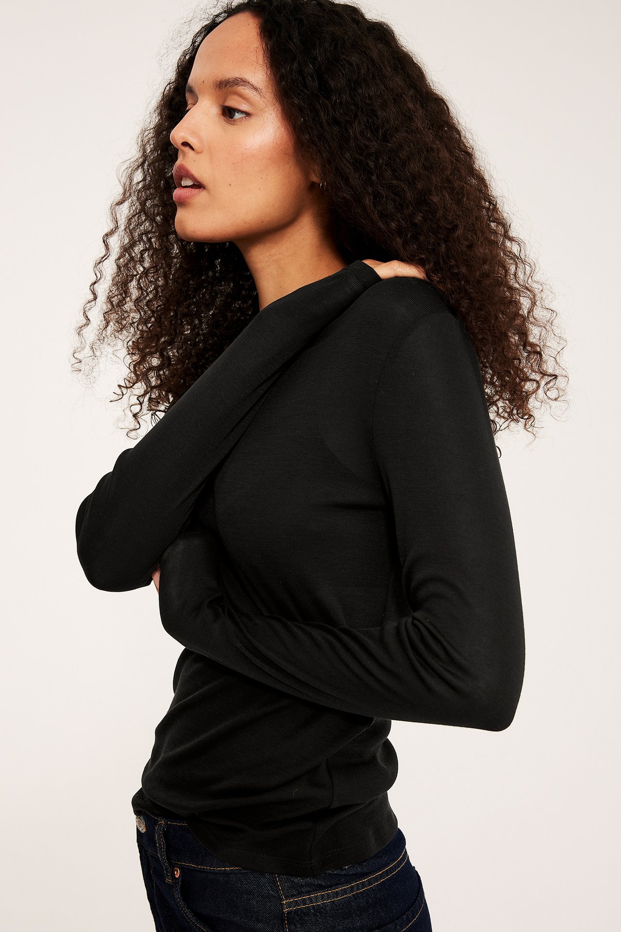 Sida vy av en kvinna i en åtsittande svart långärmad topp i lyocell och mörkblå jeans.