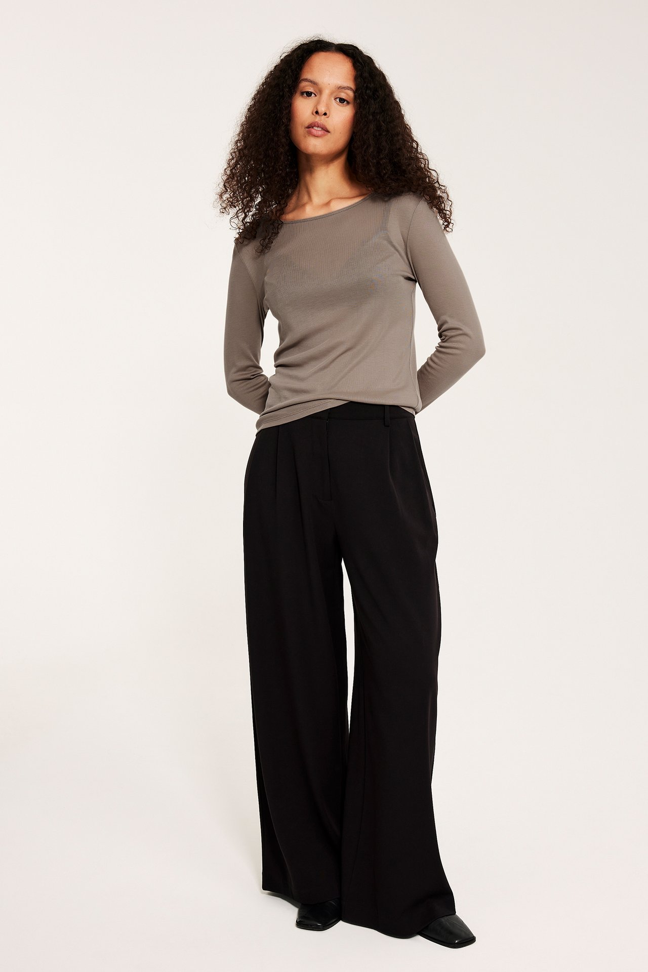 Framifrån, en kvinna i en mörkbeige långärmad topp i lyocell, svarta vida byxor och svarta platta skor.