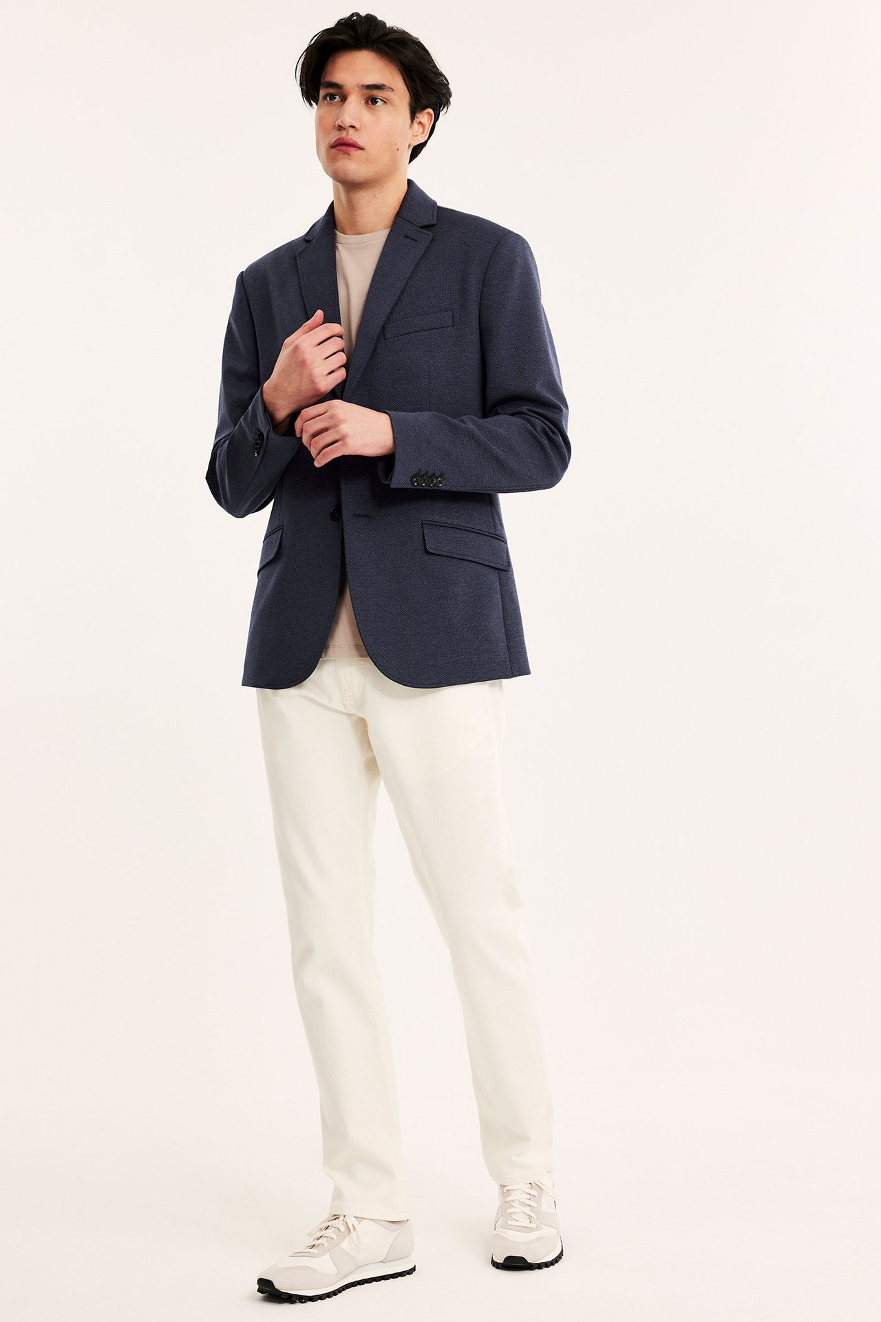 Edestä: mies tummansinisessä yksirivisessä bleiserissä, beigessä t-paidassa, kermanvärisissä housuissa ja beigissä kengissä.