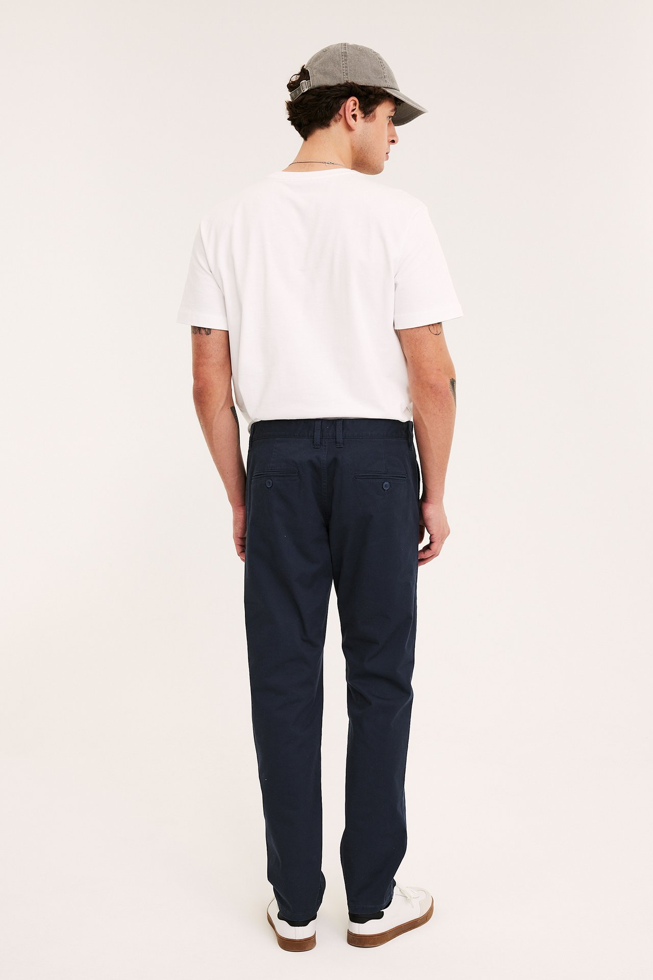 Mann bakfra i mørkeblå regular fit bomullstwillchinos, hvit t-skjorte, grå caps og hvite sneakers.