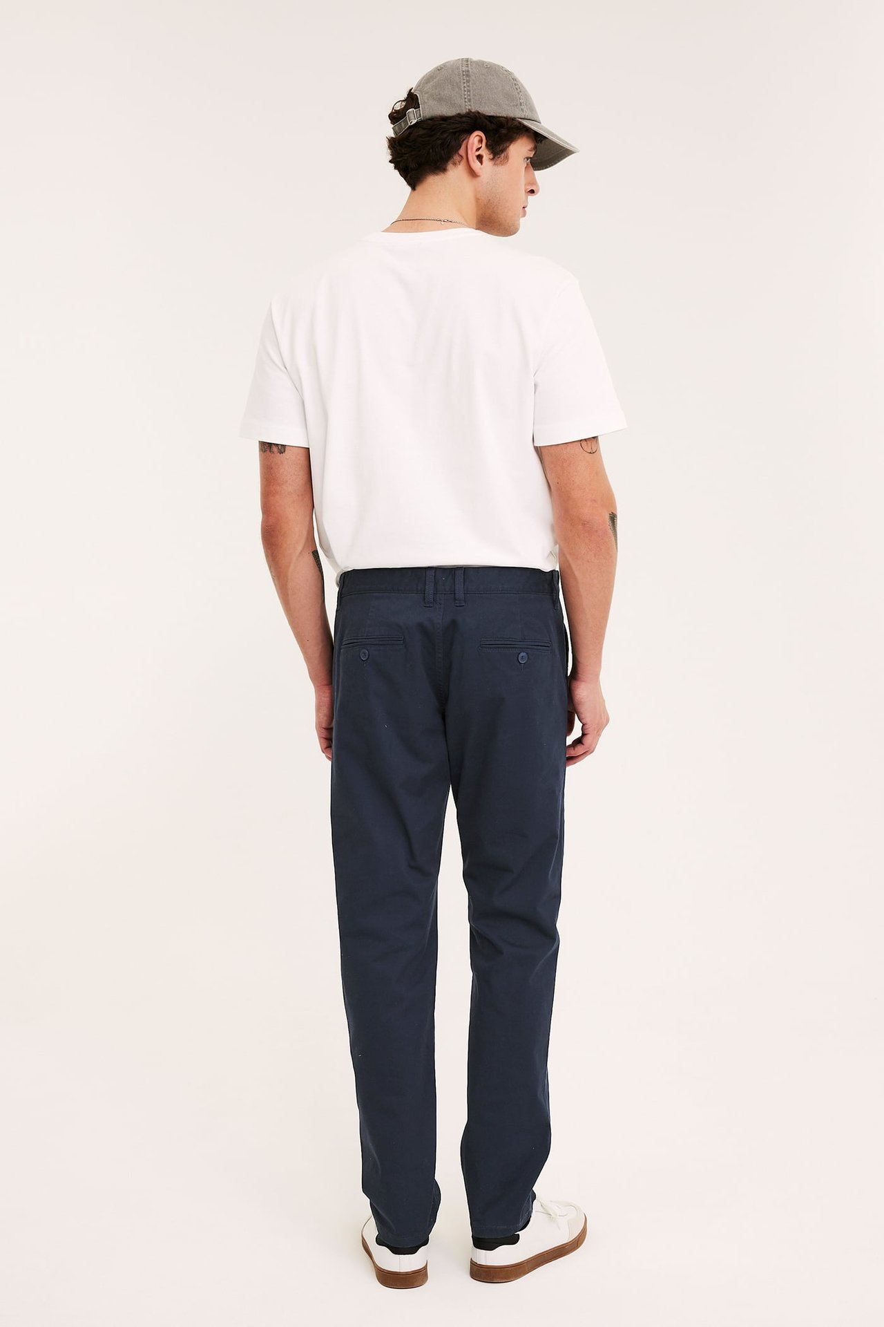 Man bakifrån i mörkblå regular fit chinos, vit t-shirt, grå keps och vita sneakers.