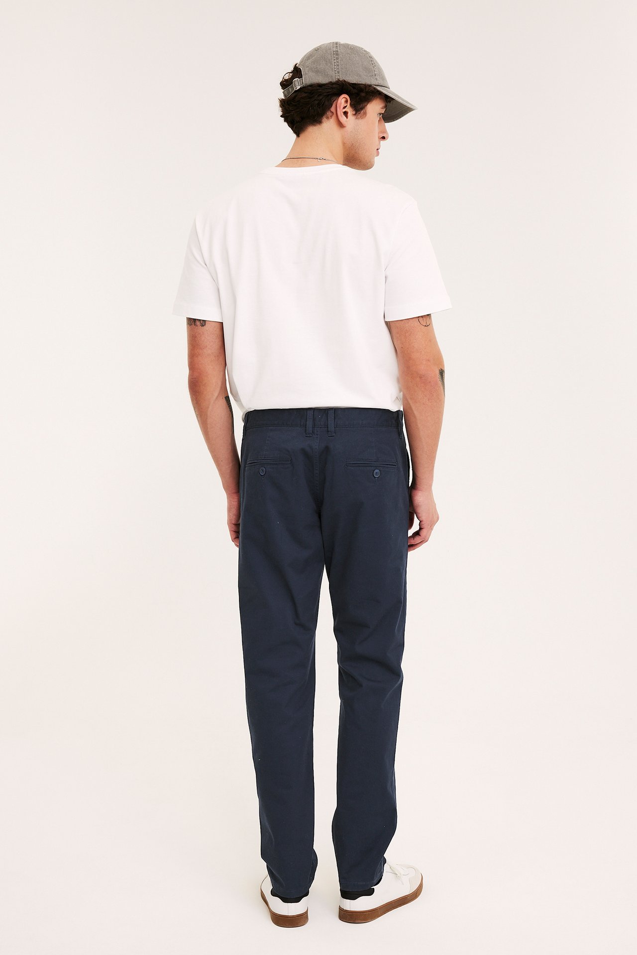 Mann sett bakfra i mørkeblå regular fit chinos, hvit t-skjorte, grå caps og hvite joggesko.