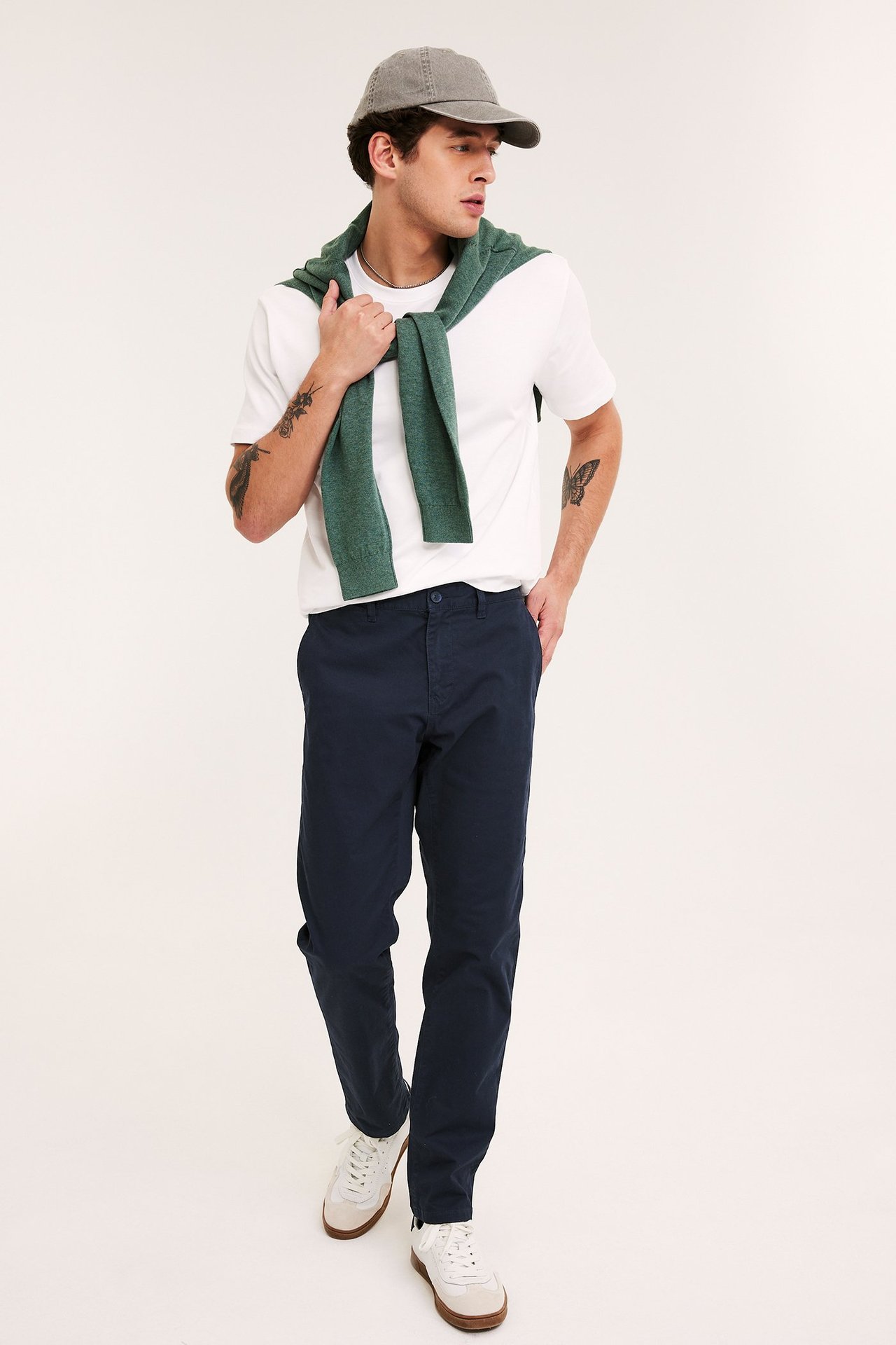 Mężczyzna w granatowych chinosach regular fit, białym t-shircie, zielonym swetrze, szarej czapce i białych trampkach.