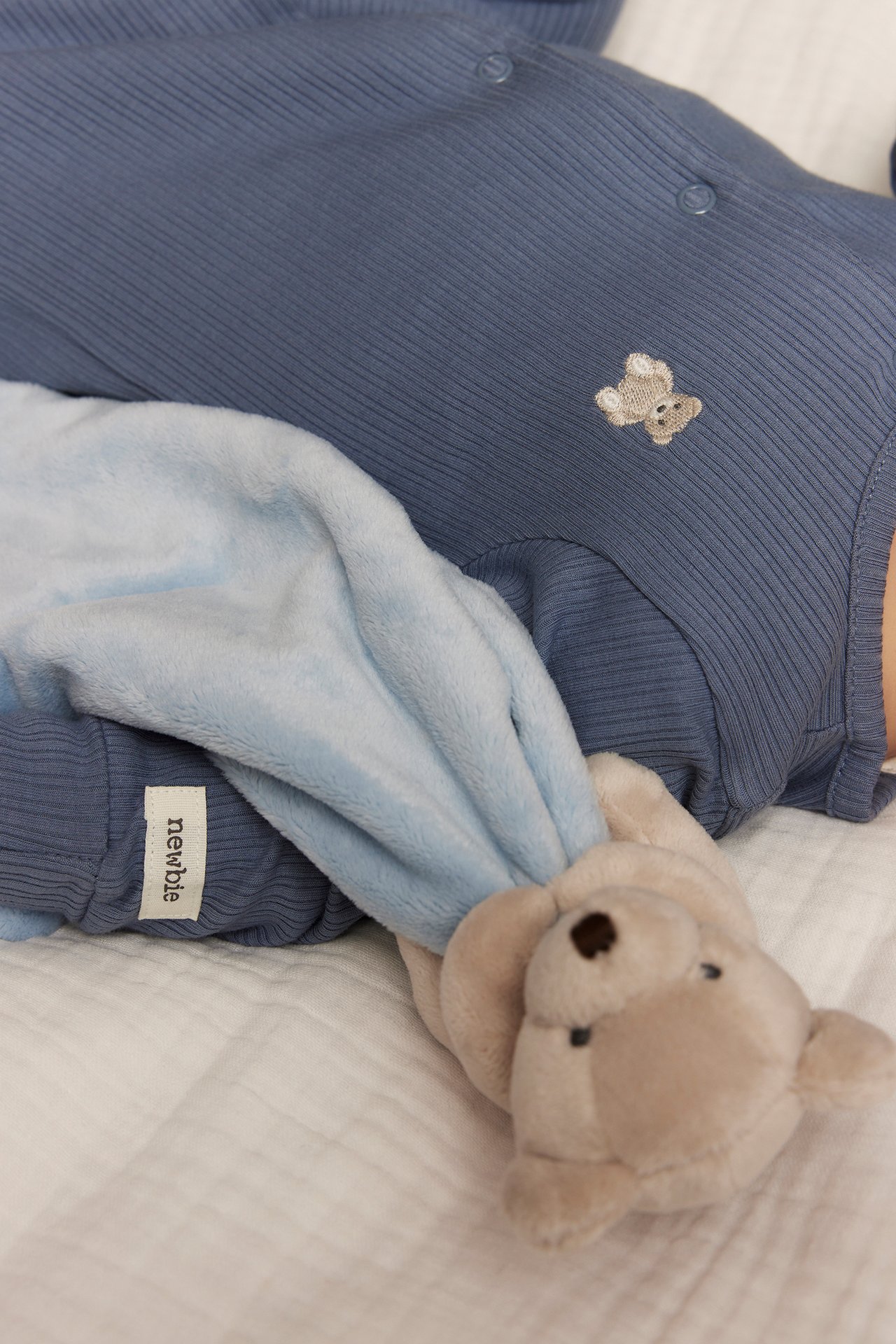 Lähikuva vauvasta sinisessä ribbipyjamassa nallekirjonnalla, pitäen vaaleansinistä peittoa ja beigeä nallea.