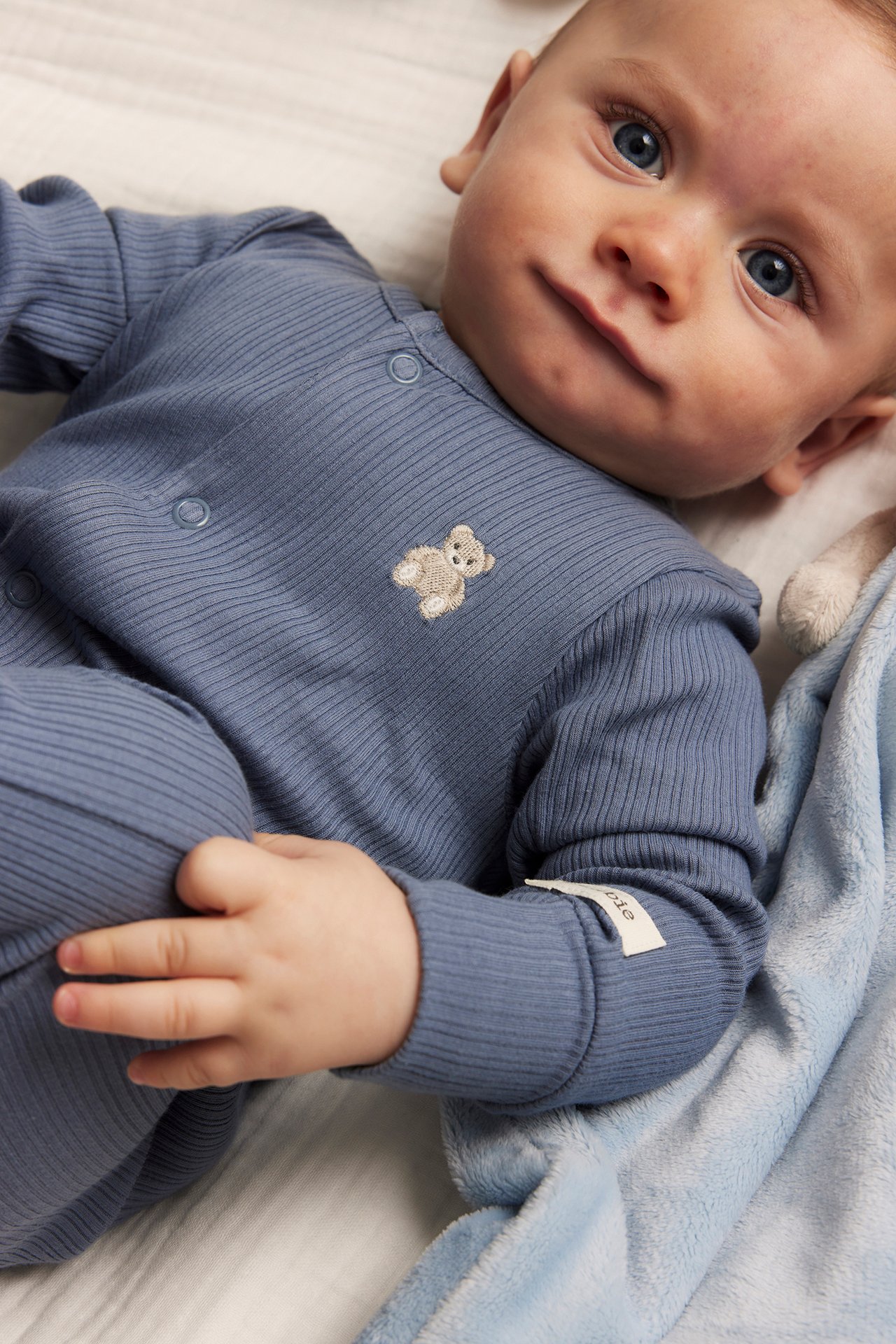 Edestä, vauva sinisessä ribbipyjamassa nallekirjonnalla. Sininen peitto näkyy.