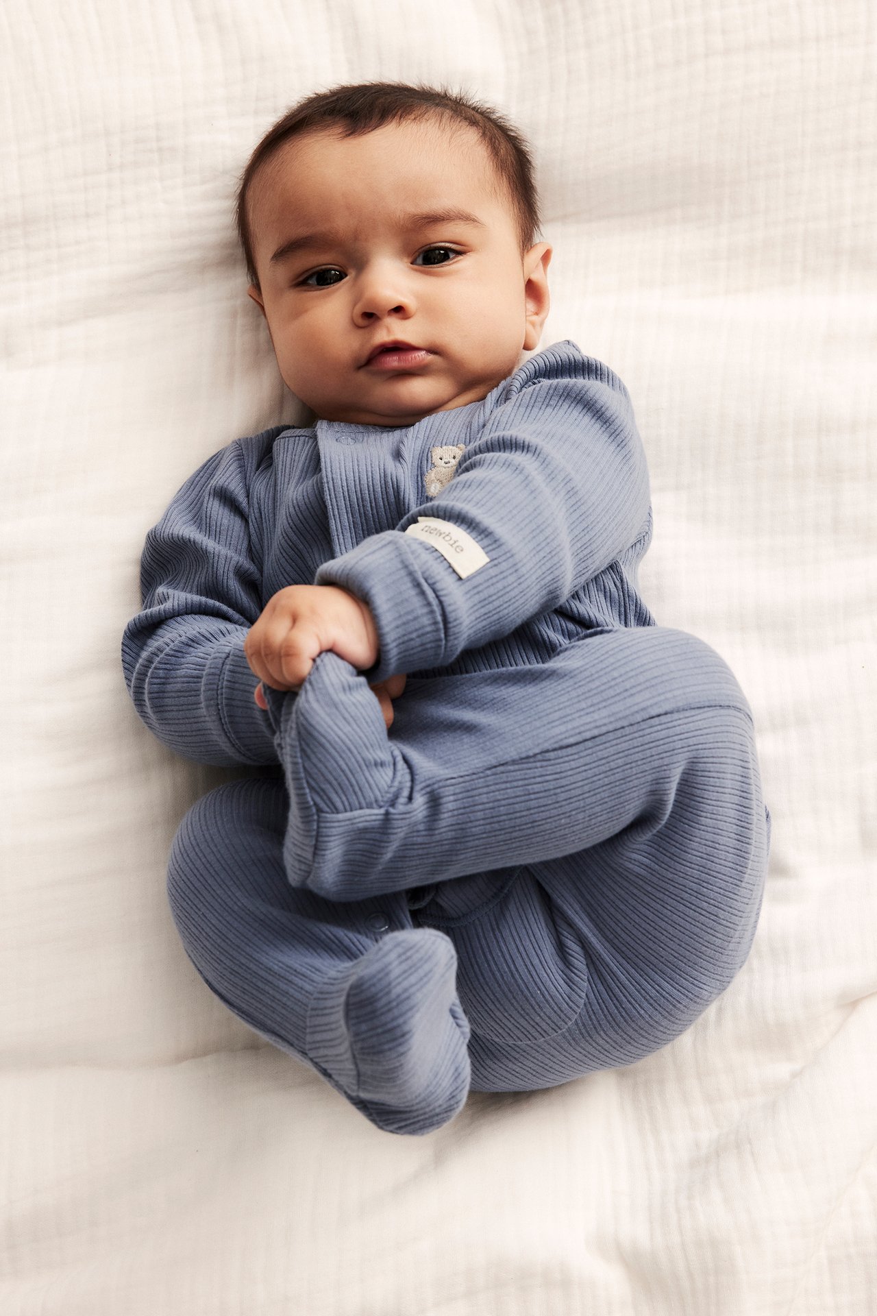 Edestäpäin, vauva yllään sininen ribattu trikoopyjama neppareilla, jaloilla ja nallekirjonnalla.