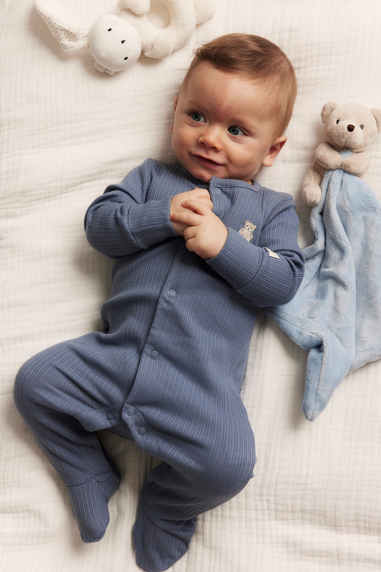 Ylhäältä, vauva sinisessä ribbipyjamassa neppareilla ja nallekirjonnalla, sinisen uniriepun vieressä.