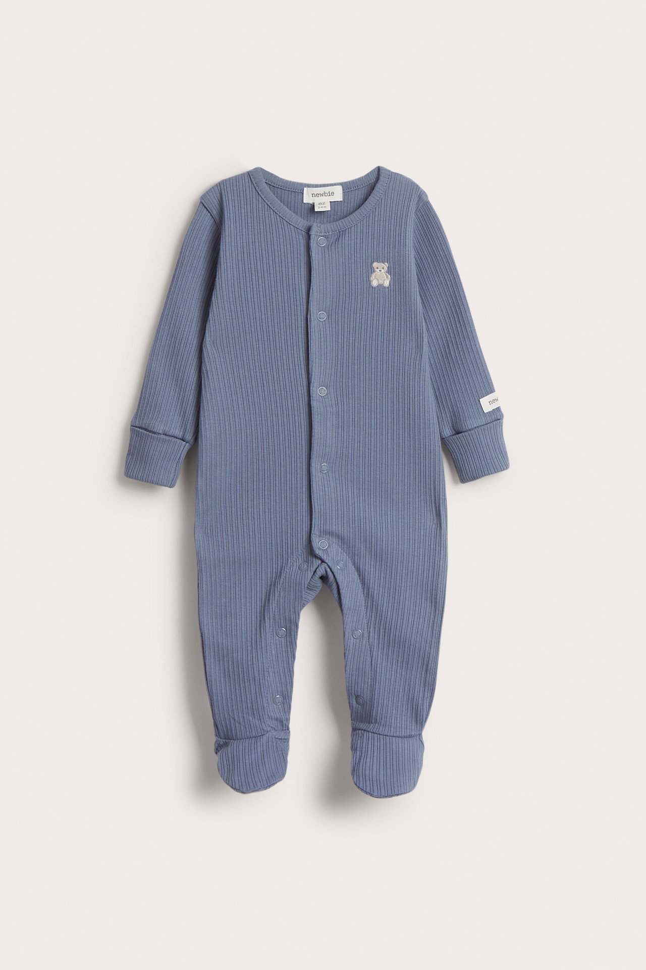 Edestä näkymä sinisestä ribatusta vauvan pyjamasta, jossa nepparit, integroidut jalkaosat ja pieni nallekirjailu.