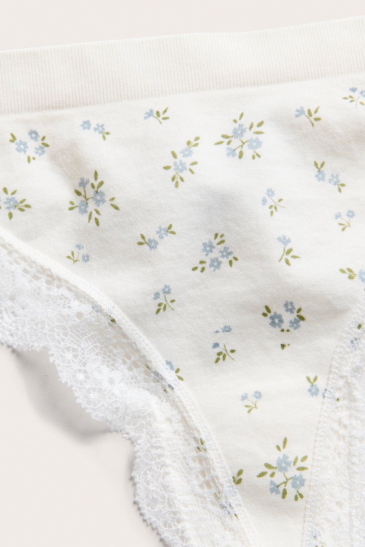 Zbliżenie na kremowe stringi z drobnym niebieskim wzorem kwiatowym i białą koronką.