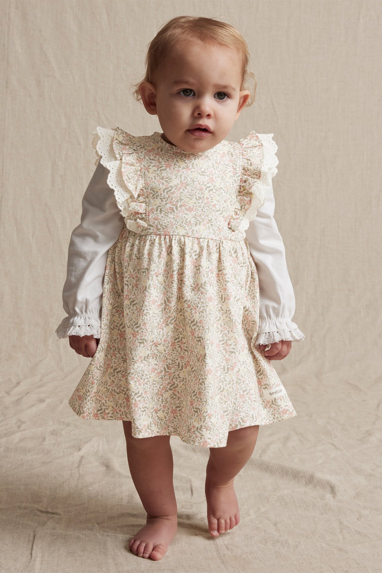 Frontbilde av en baby i en offwhite blomstret kjole med volanger over en hvit langermet topp.