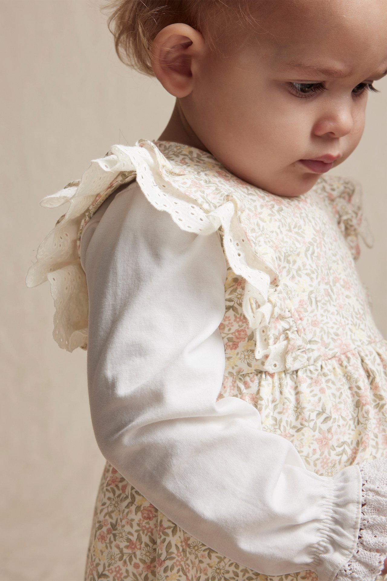 Nærbilde sidevisning av baby i offwhite blomsterkjole med blondevolanger over en hvit langermet topp.