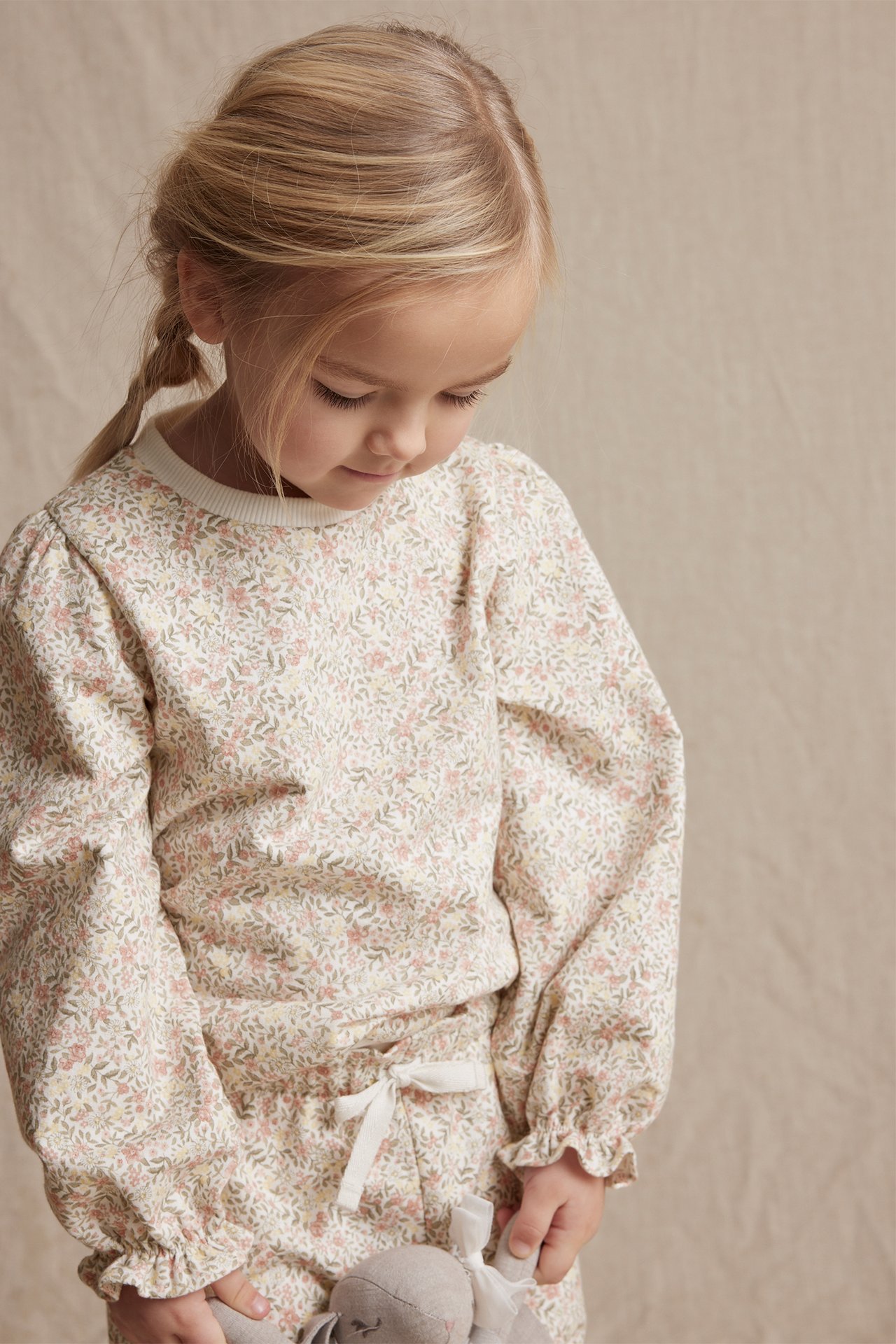 Frontvisning: Barn i offwhite blomstret bomullssweatshirt med puffermer, matchende bukser, holder en grå leke.