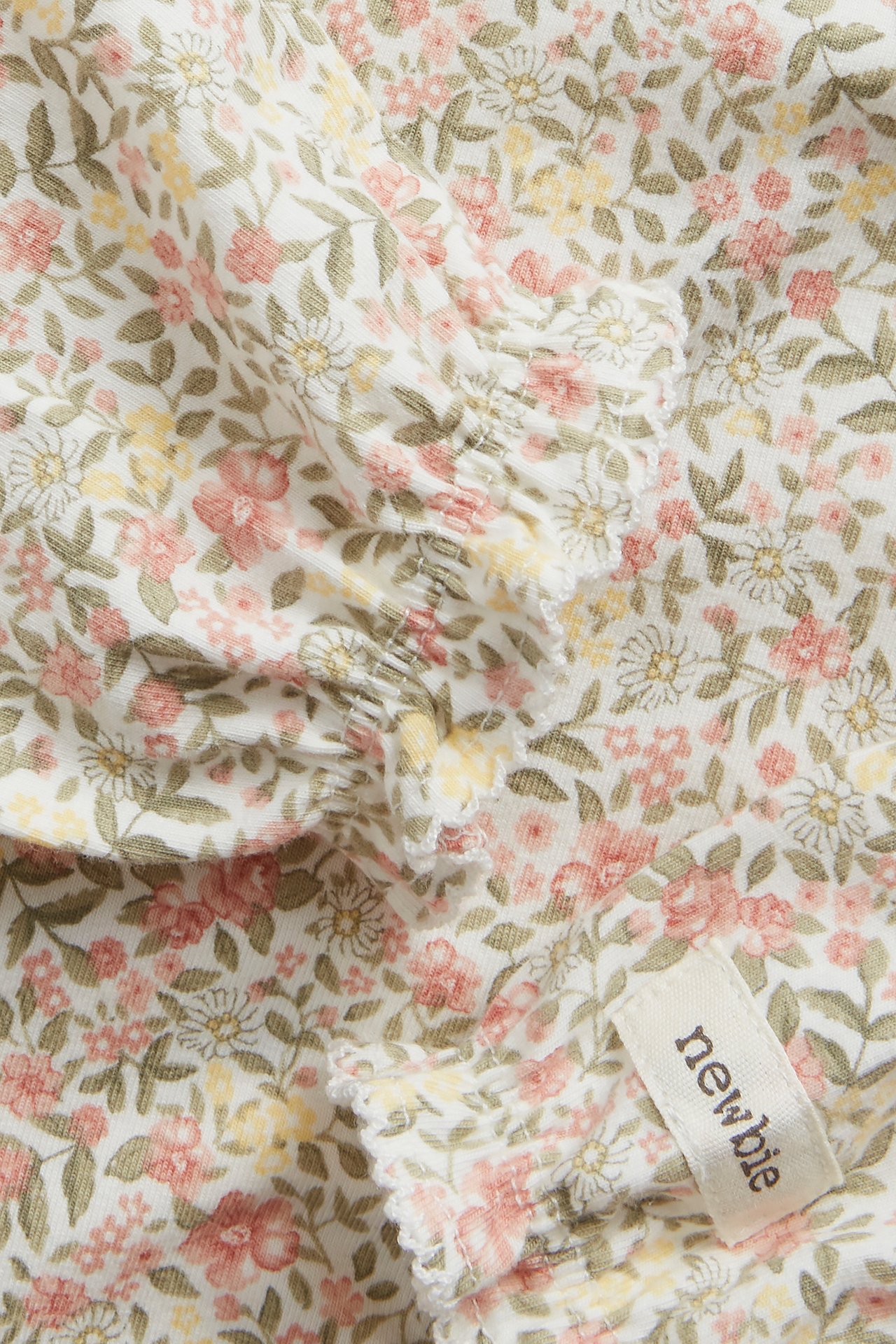 Nærbilde av en offwhite babybody i jersey med lang erm, rosa/gult blomstermønster og volangermer.