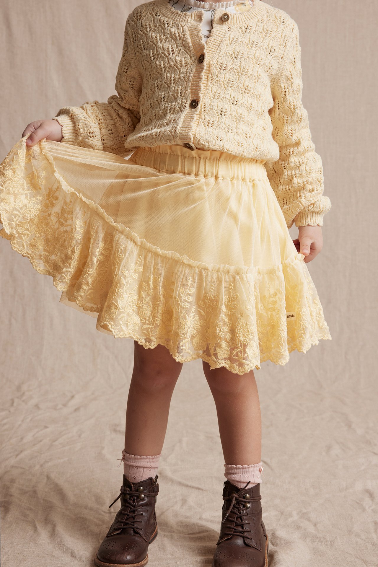Frontvisning: Barn i gult meshskjørt med brodert volang, kremfarget strikket cardigan, hvit topp og brune støvler.