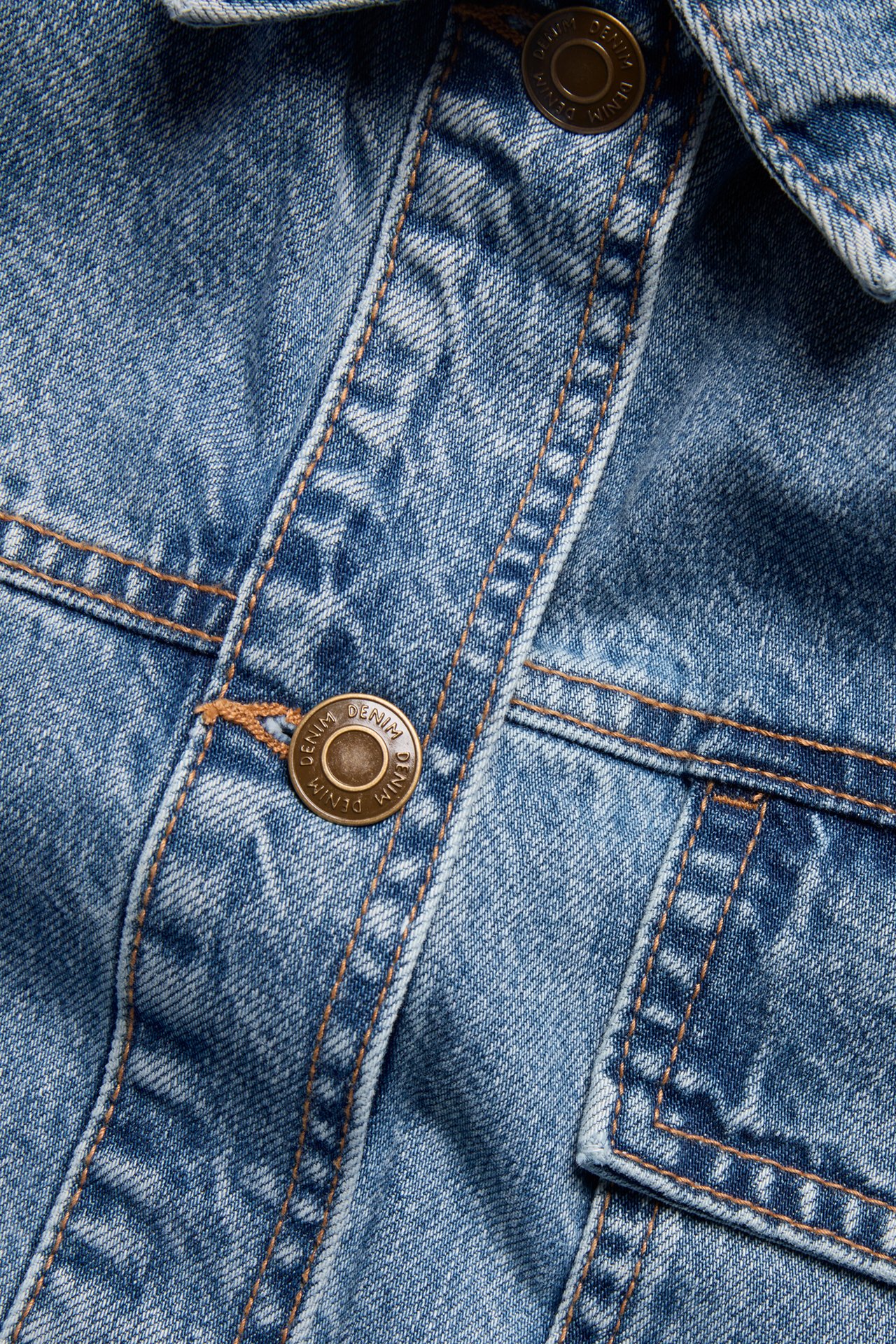 Närbild av en blå denim shacket med en bronsfärgad knapp, knappslå och bröstficka.