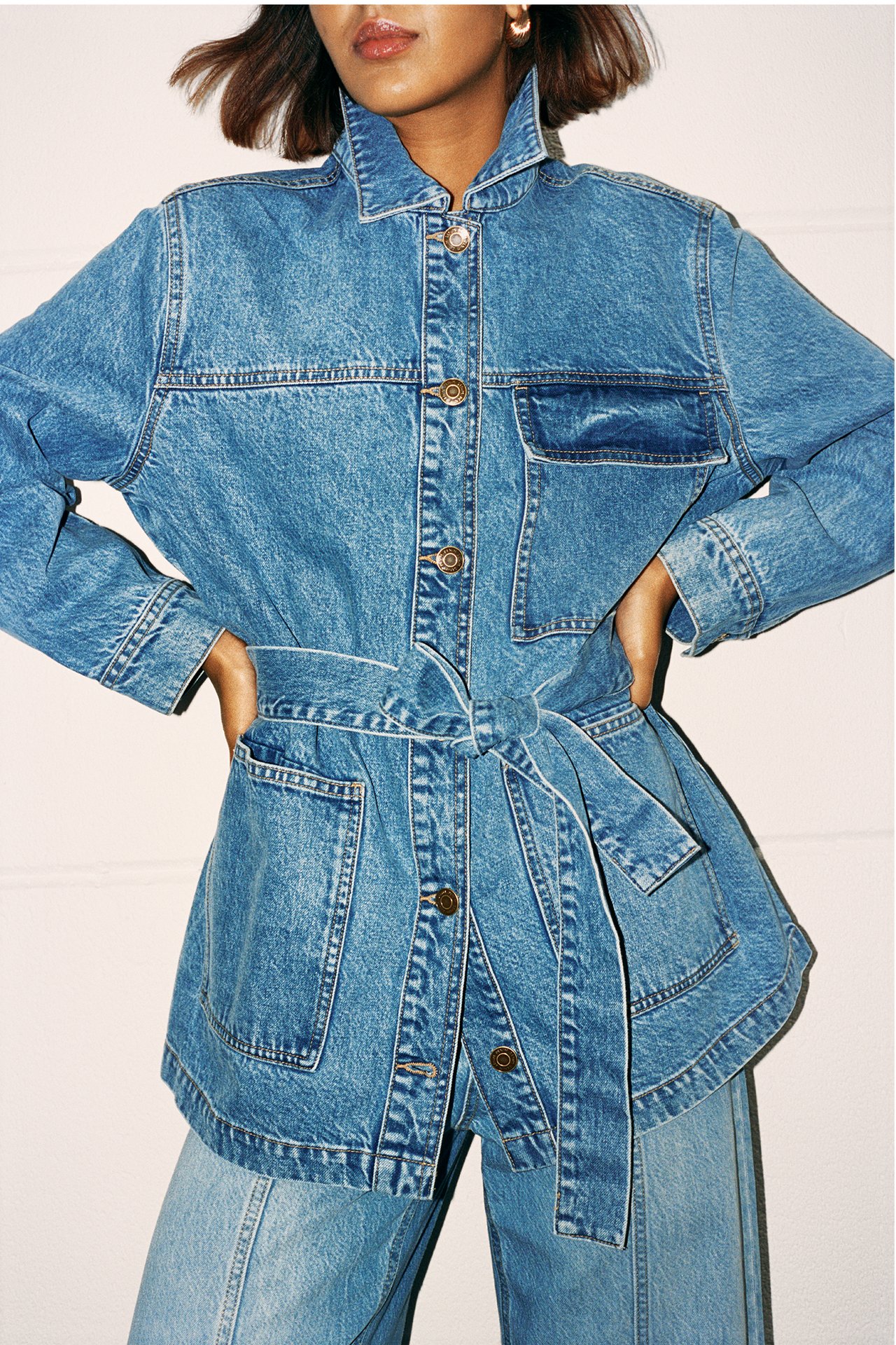 Framifrån: kvinna i blå jeansjacka med bröstfickor, knutet bälte och matchande jeans.