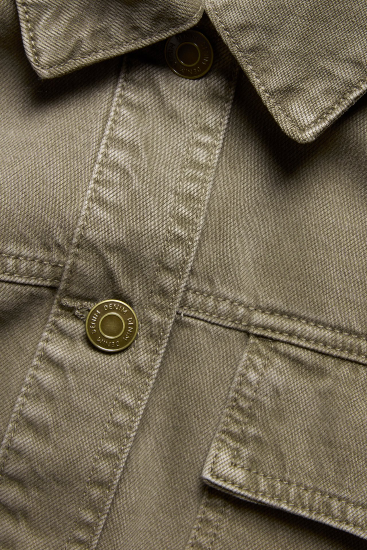 Nærbilde av en grønn bomullsdenim utility shacket med krage, messingknapper og synlige sømmer.