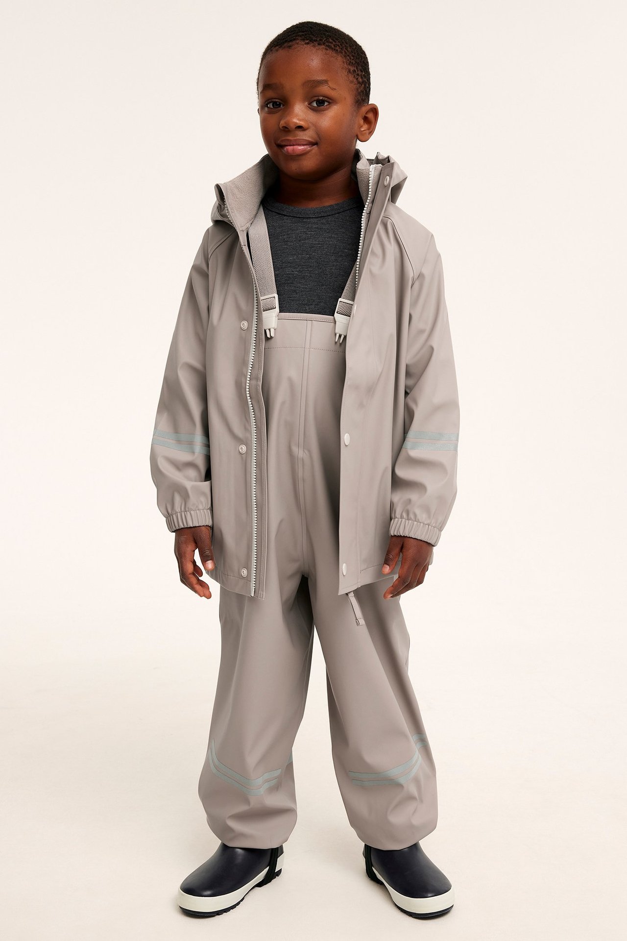 Edestä: Lapsi beigessä sadeasussa heijastinnauhoilla, tummanharmaassa t-paidassa ja sinisissä kumisaappaissa.