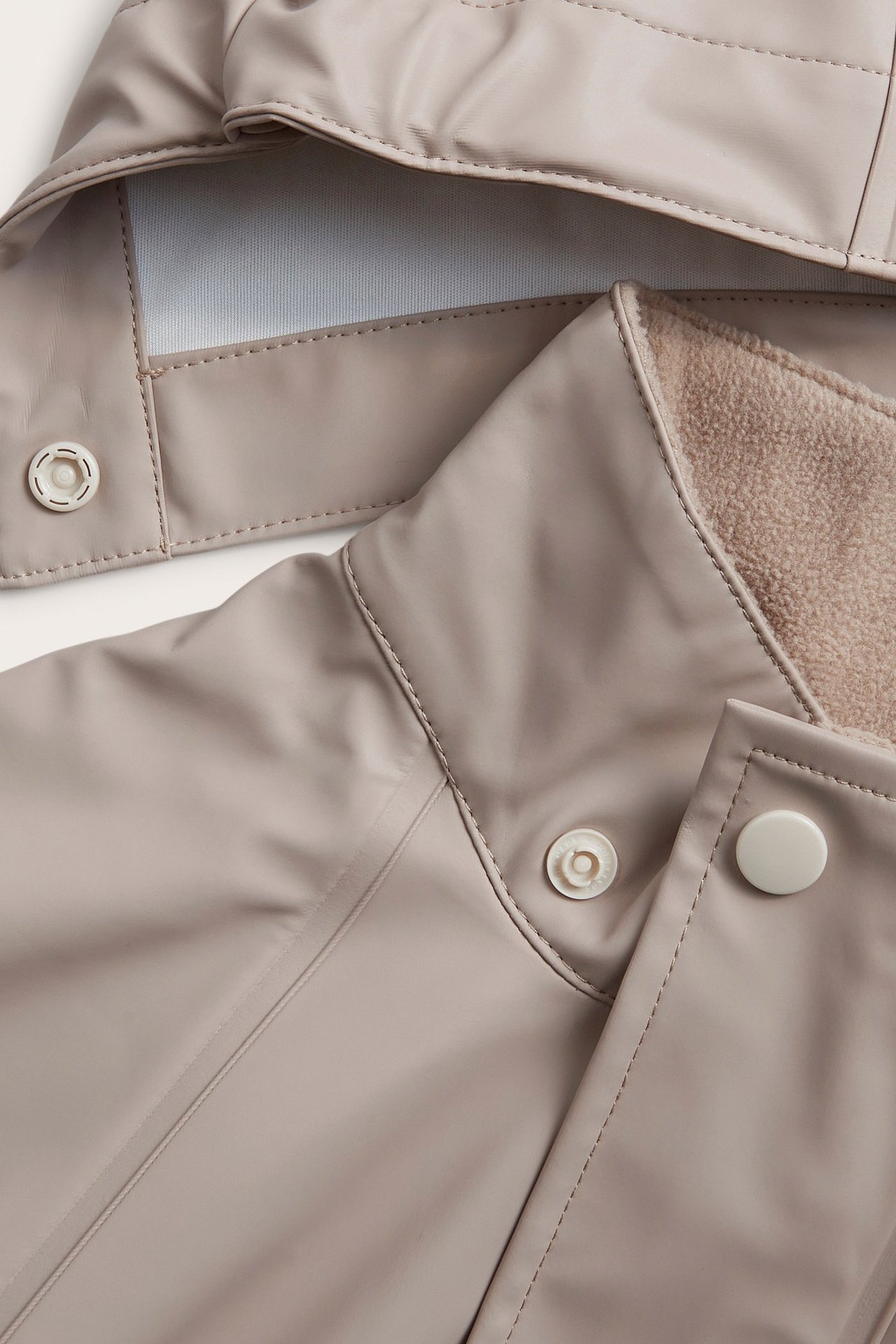 Zbliżenie beżowej kurtki przeciwdeszczowej dla dziecka z zatrzaskami i widoczną polarową podszewką przy kołnierzu.