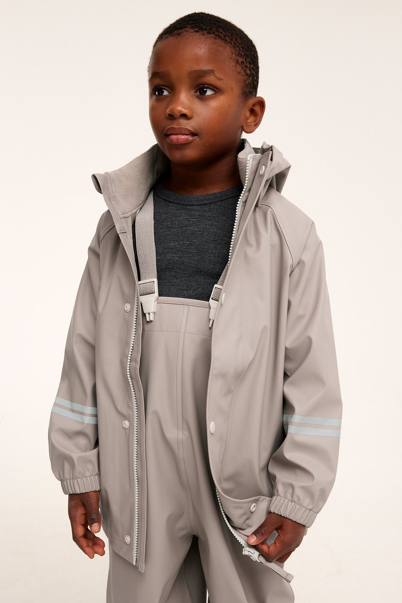 Barn bär en beige regnjacka med avtagbar luva, över matchande regnbyxor och en mörkgrå t-shirt.