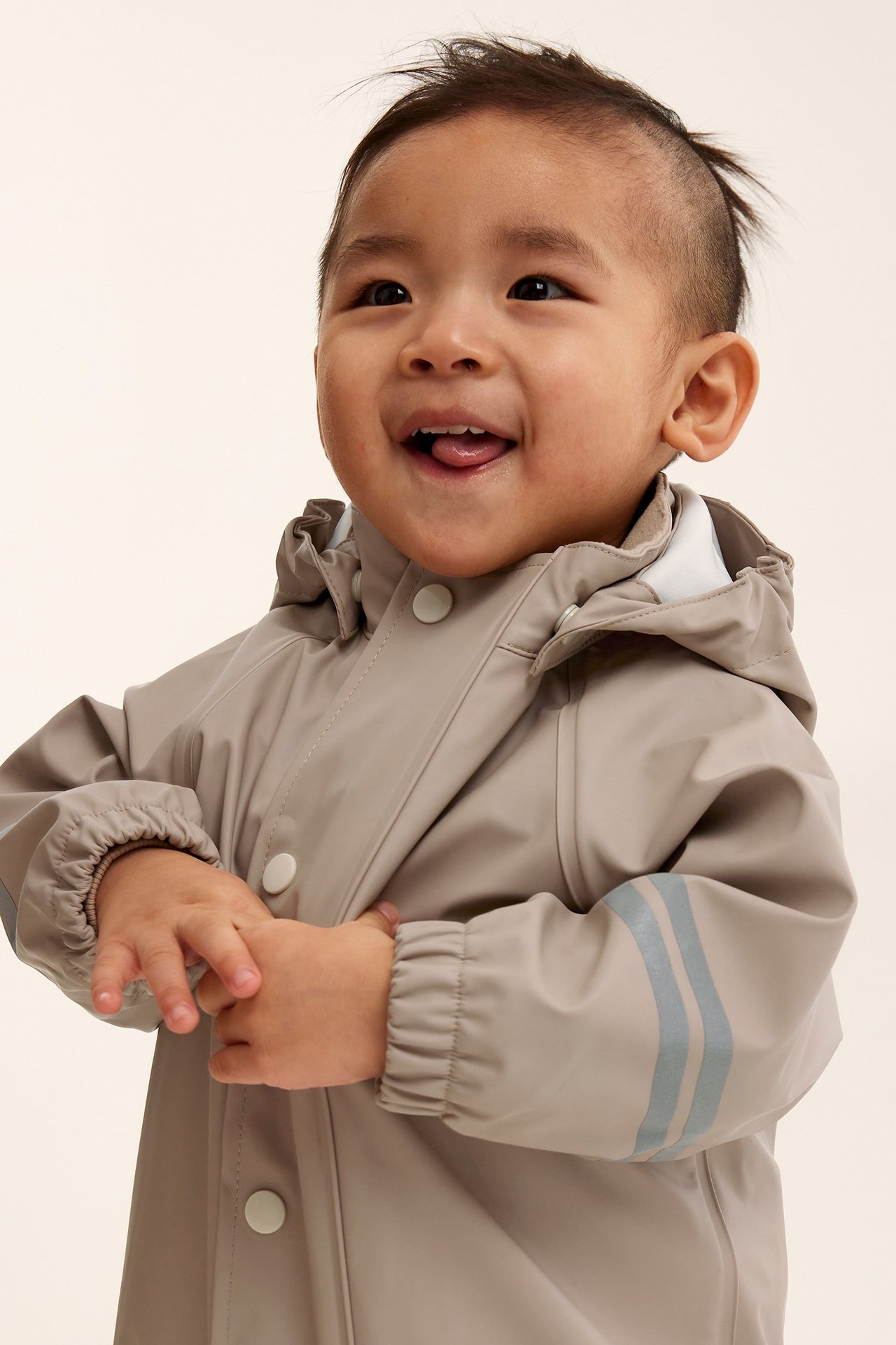 Frontvisning av baby i beige regnjakke med hette, knappestolpe og striper.